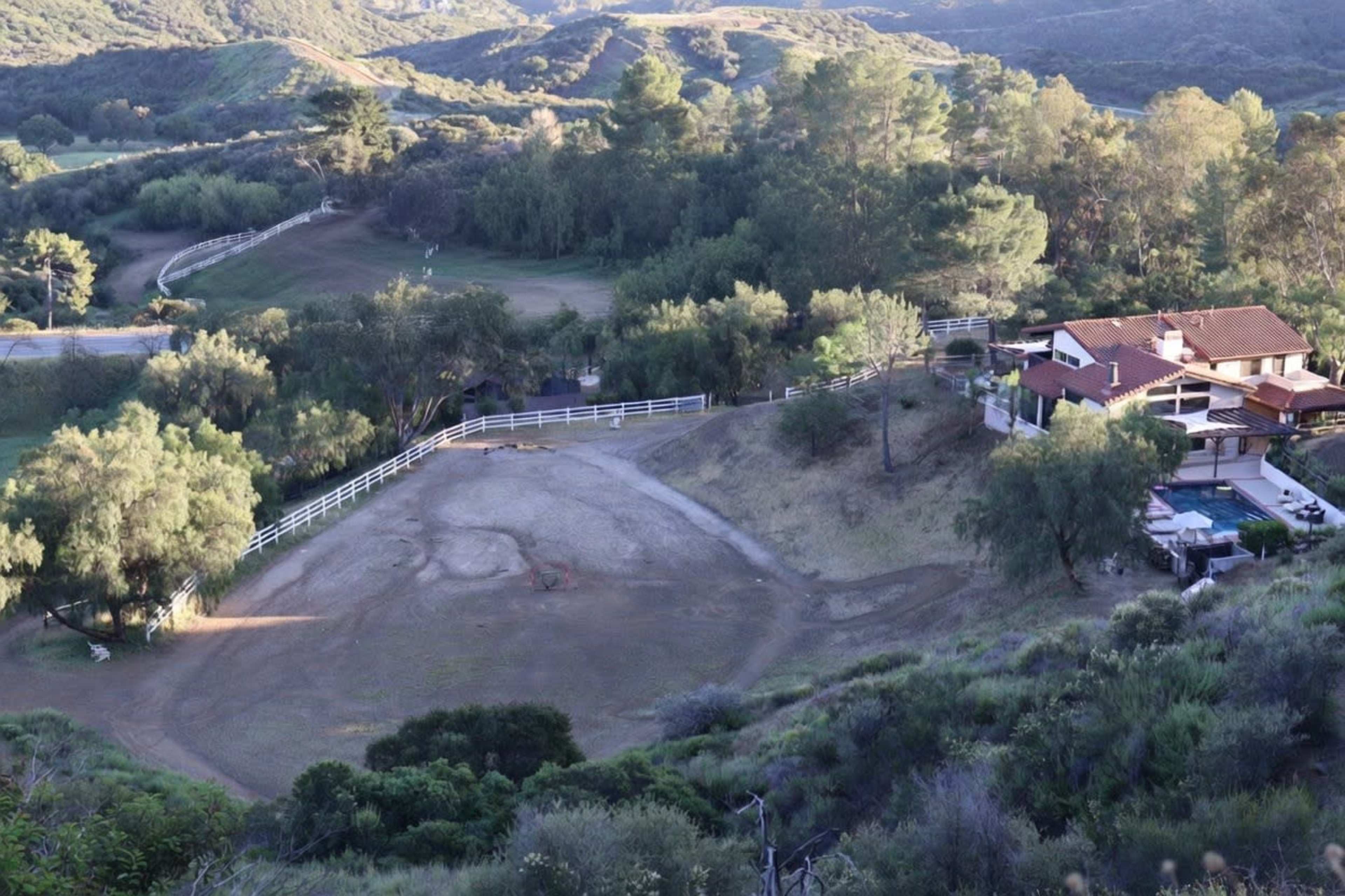 The image shows a hillside property with a house and an empty horse riding area surrounded by trees and rolling hills.