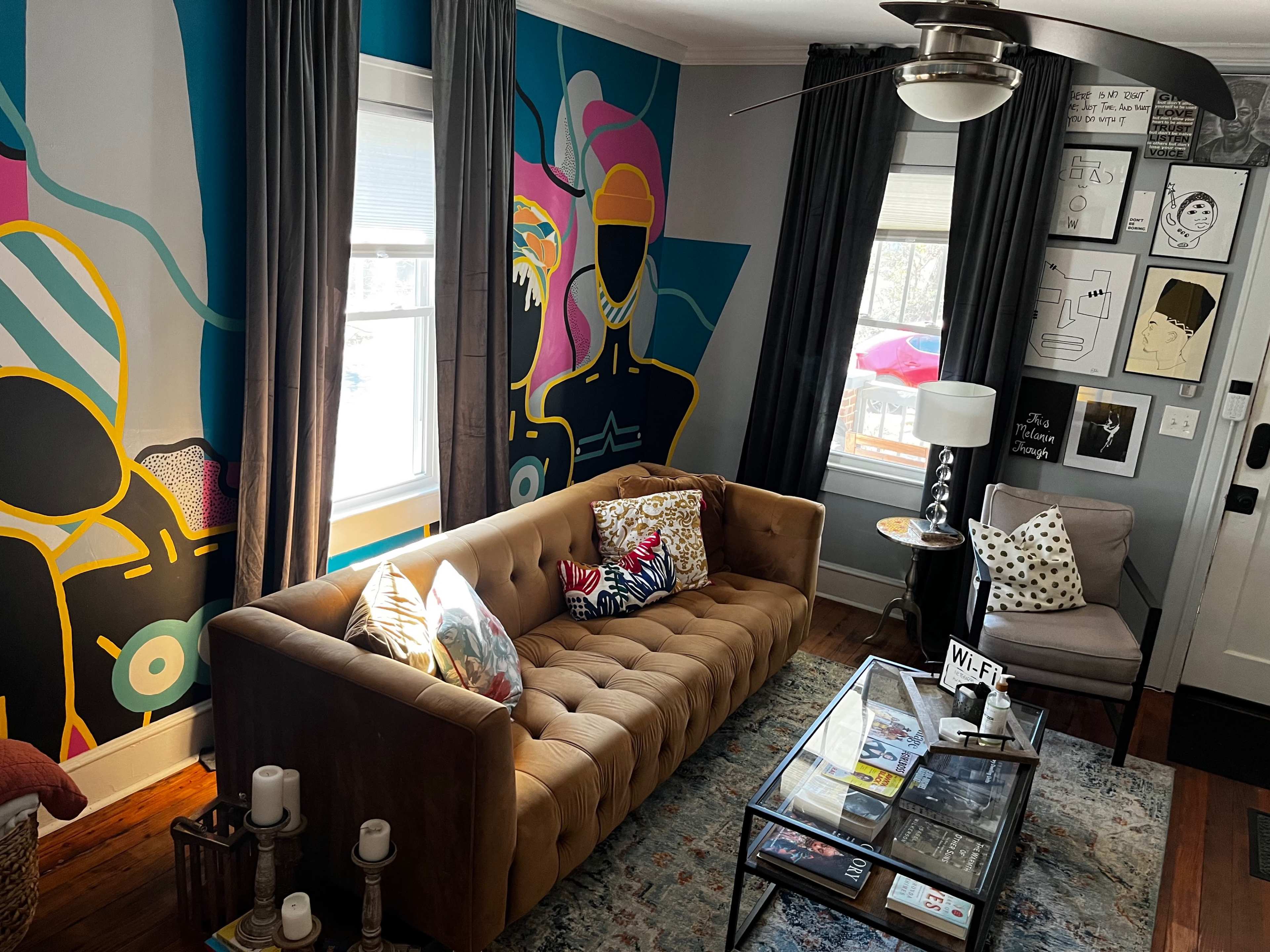 The room features a tan sofa with decorative pillows, a glass coffee table, and colorful wall art, accompanied by large windows adorned with gray curtains.