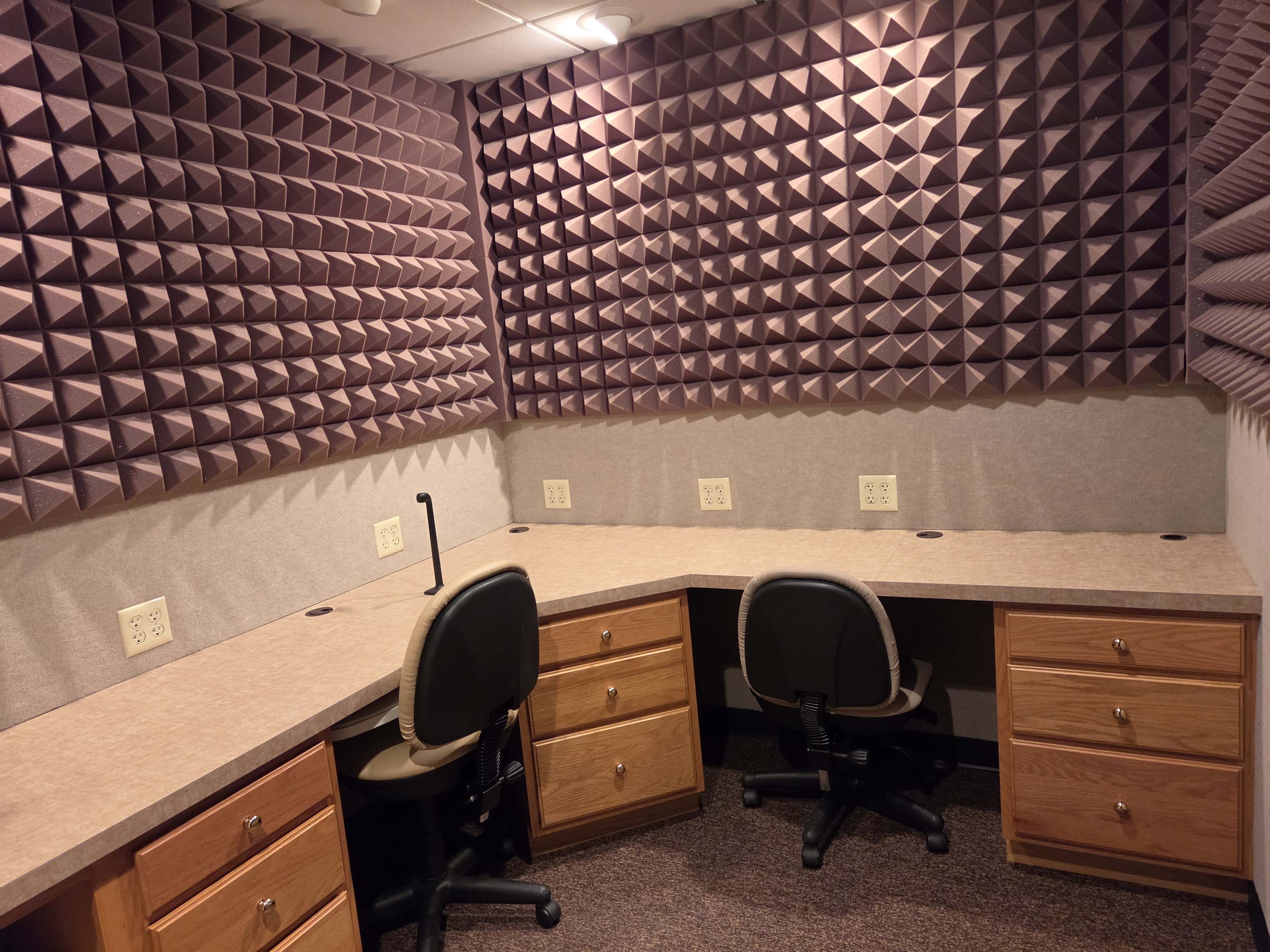 The image shows a small soundproof recording studio with textured walls, two office chairs, and a curved wooden countertop with electrical outlets.