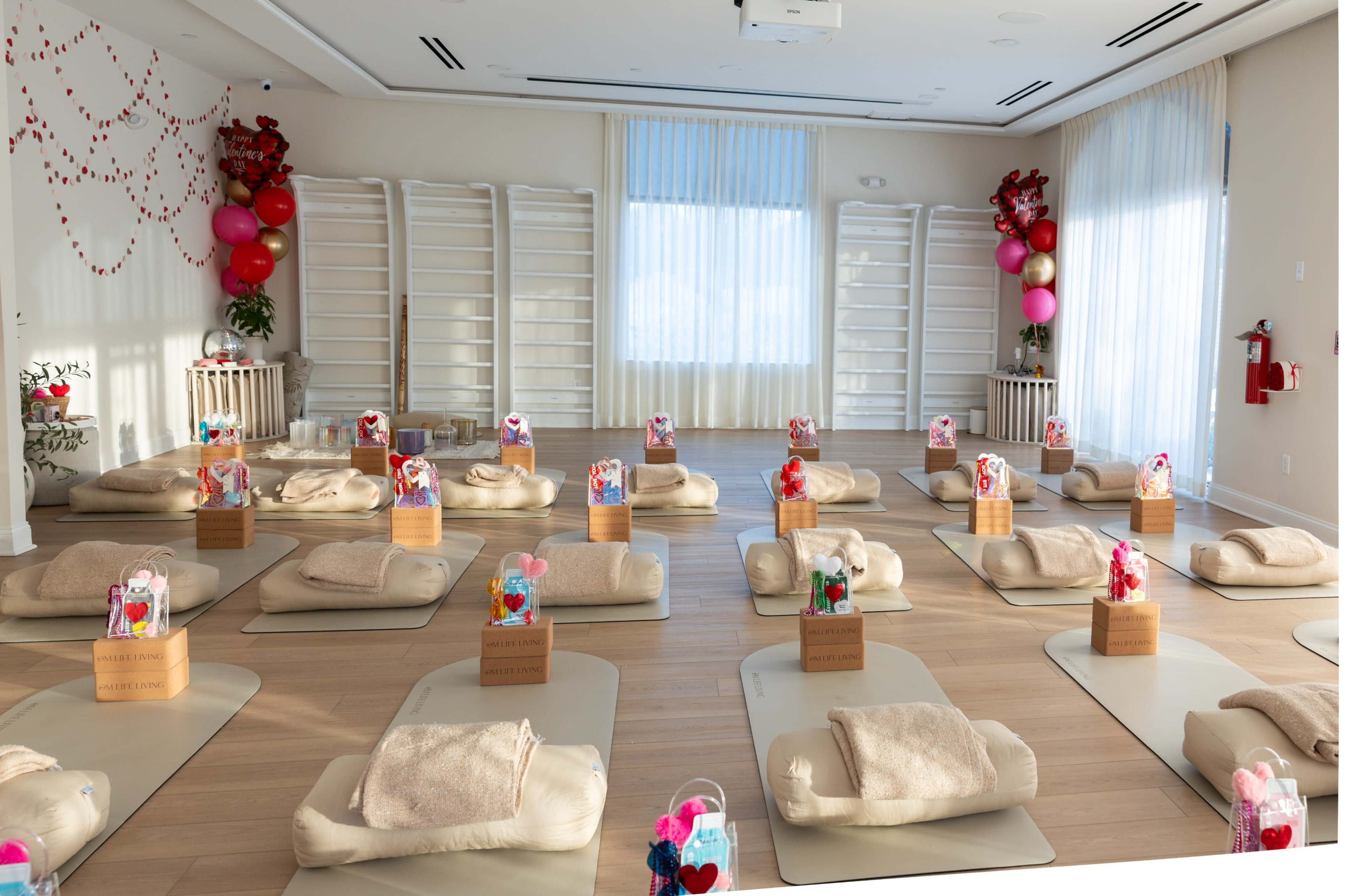 The image shows a yoga or meditation studio arranged with beige mats, each featuring a gift basket, in a bright room with large windows and decorative balloons.