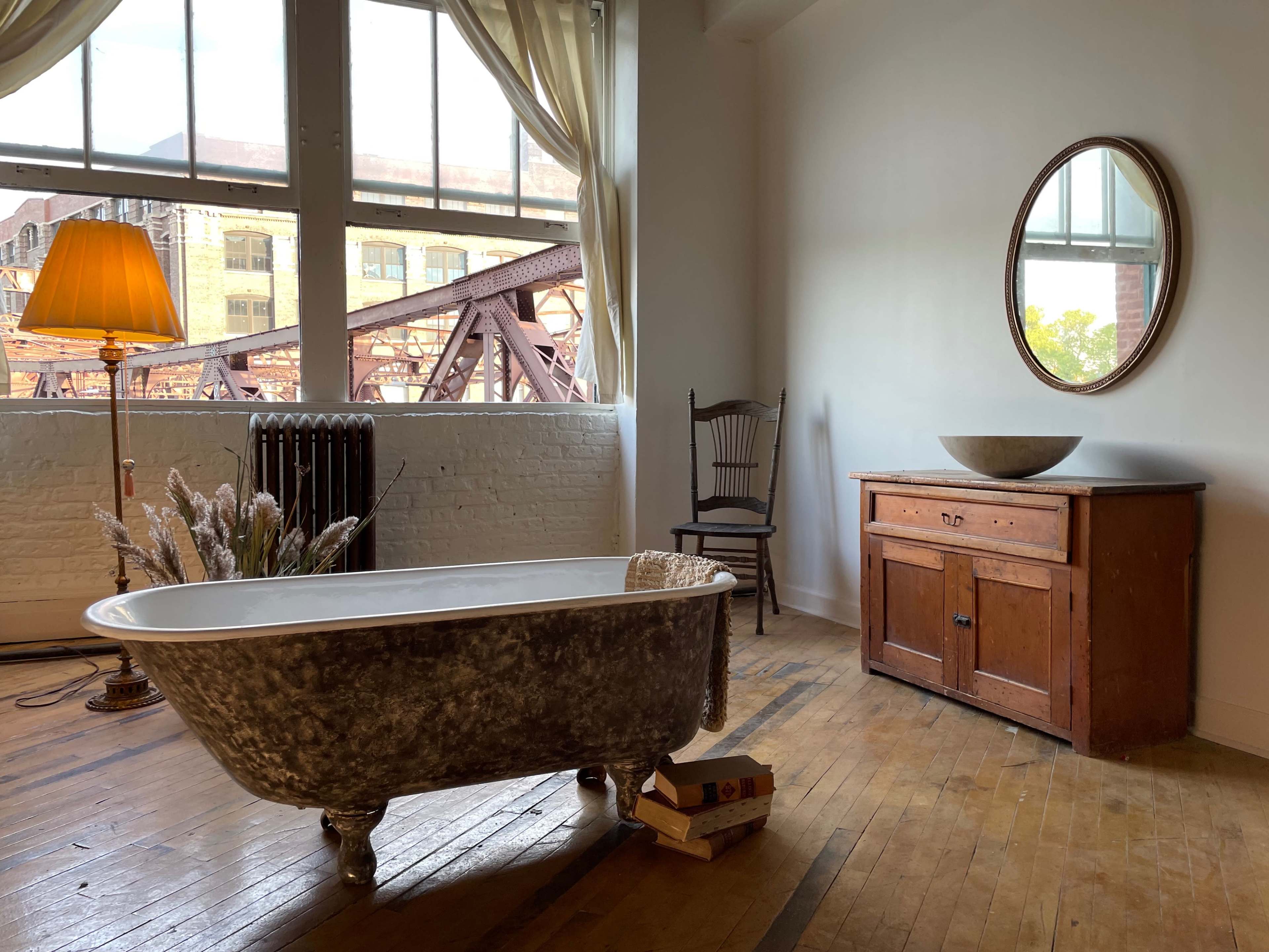 The image shows a bright room featuring a freestanding bathtub, a wooden dresser, a chair, and a lamp, with large windows revealing a view of a bridge.