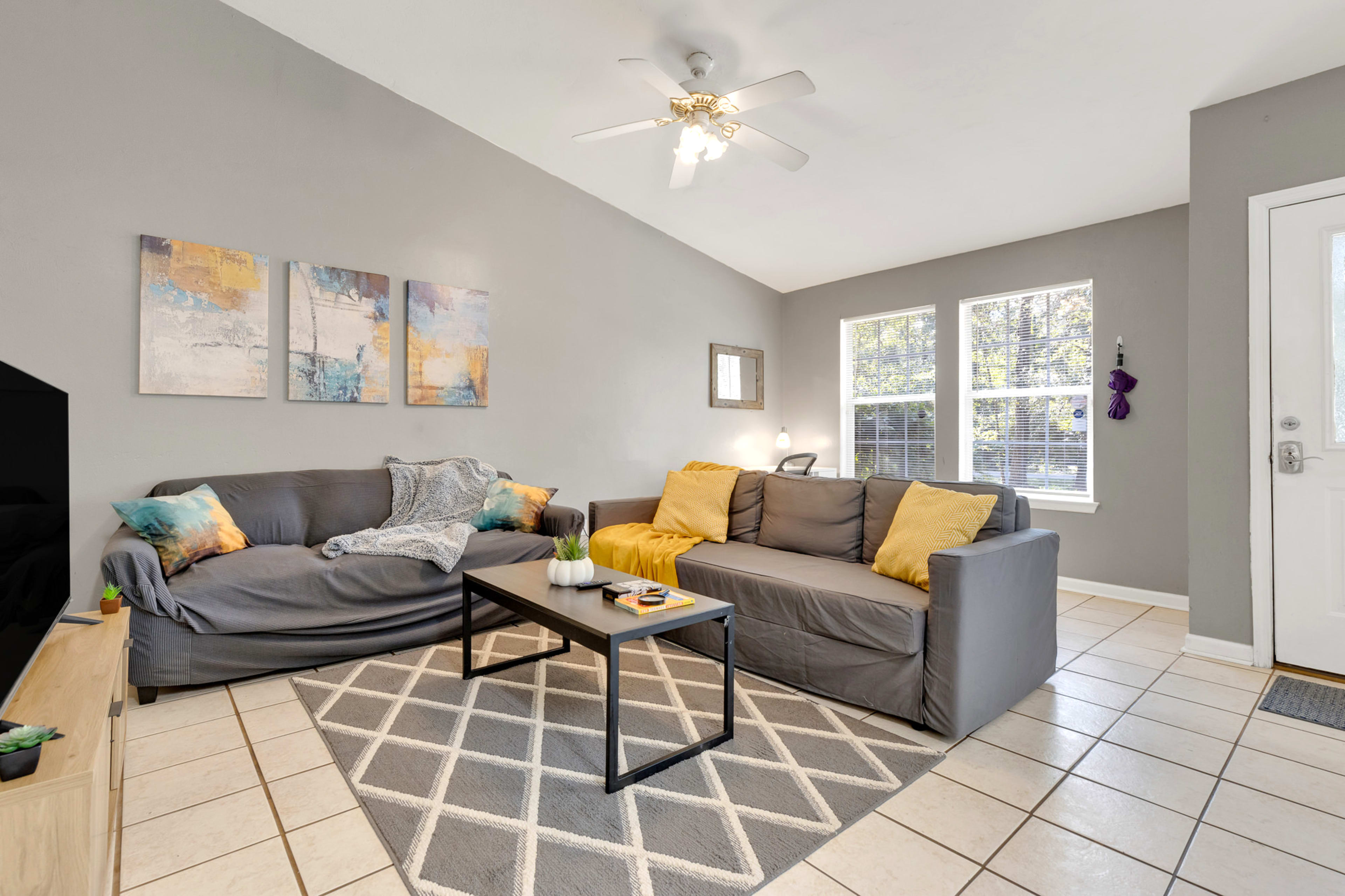 The living room features two gray sofas with yellow pillows, a coffee table on a patterned rug, and large windows allowing natural light.