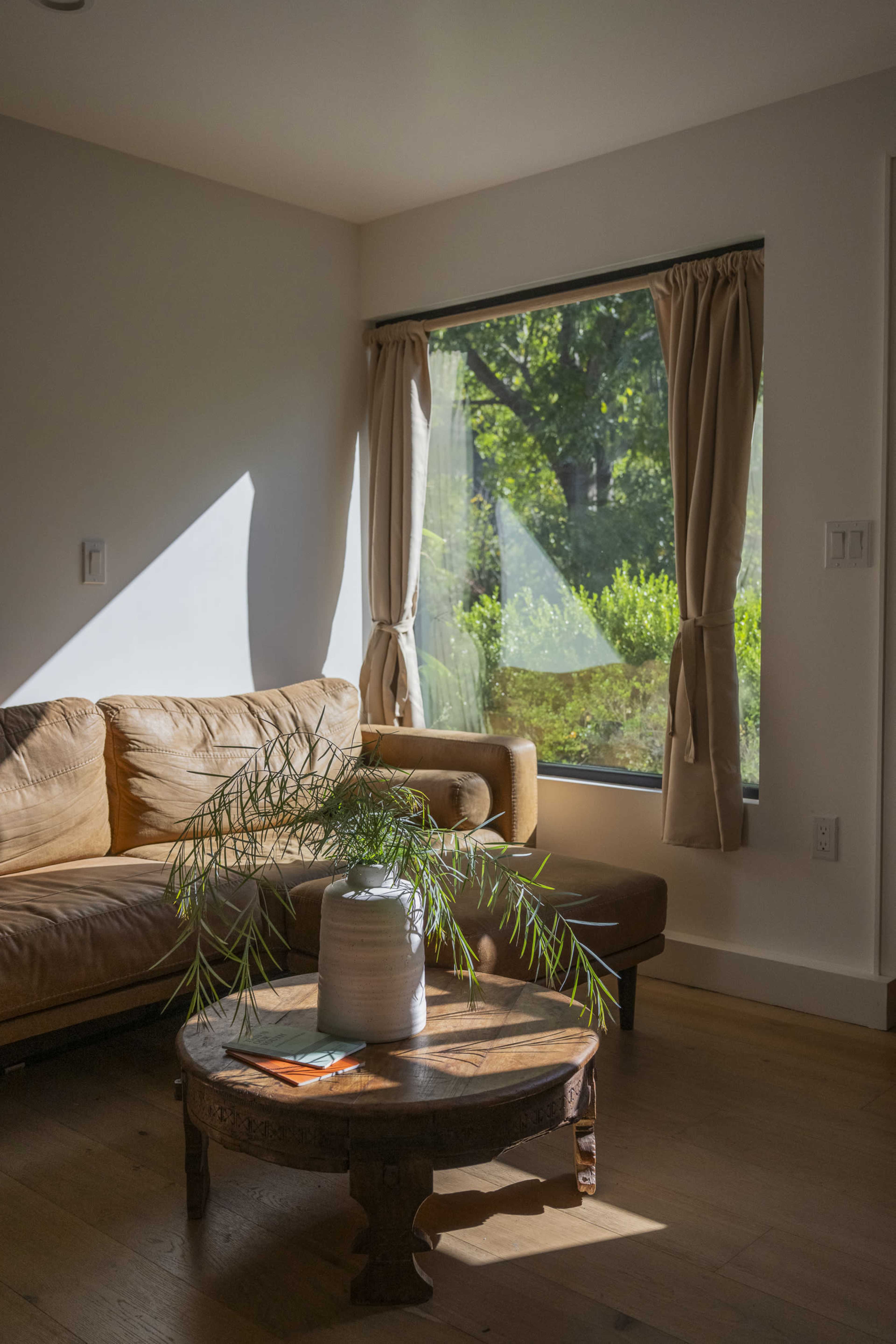 A cozy living room features a brown leather sofa and a circular wooden table, with a large window showcasing greenery outside.