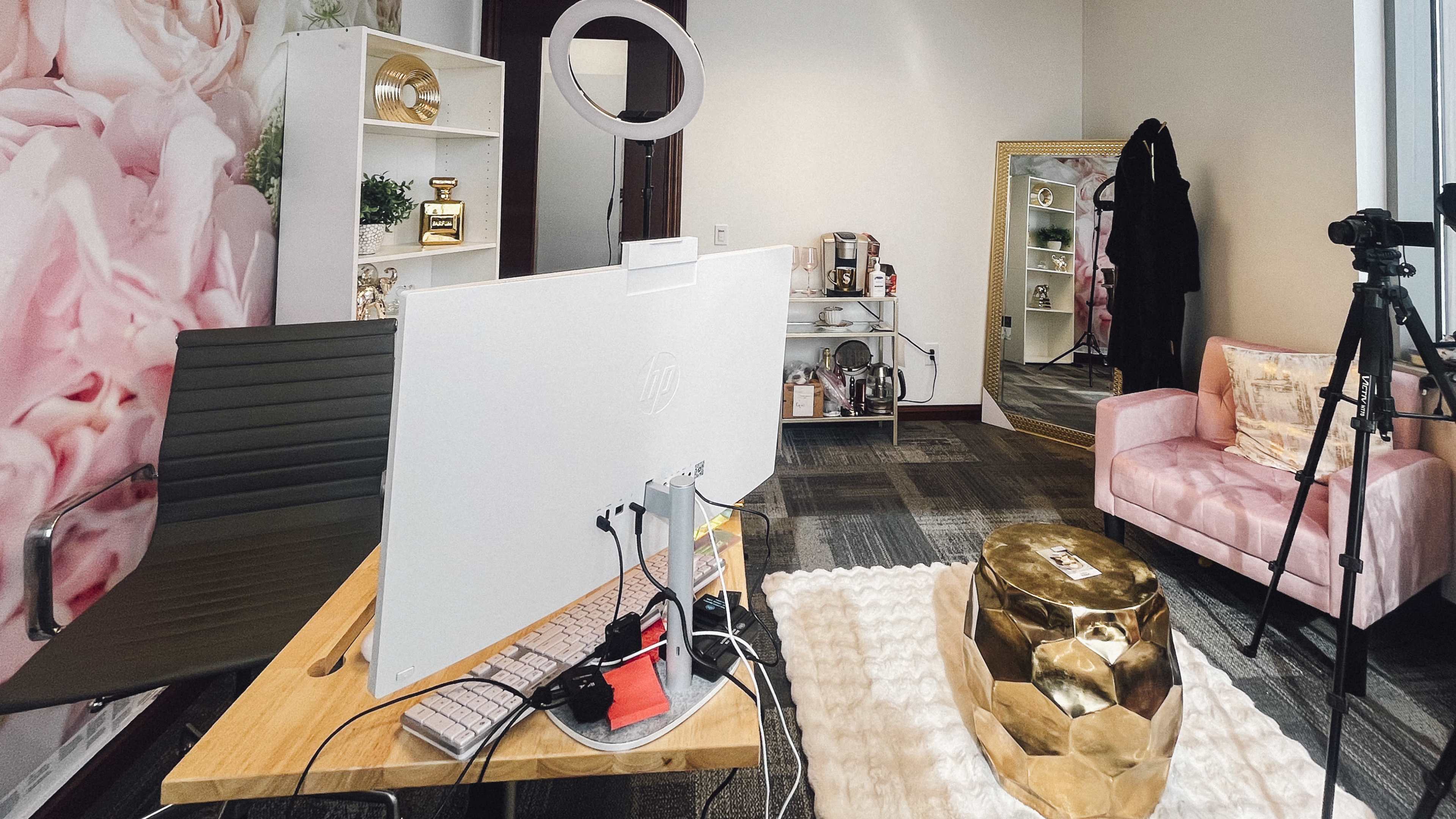 The image shows a well-arranged office space featuring a large computer on a desk, a pink chair, a mirrored cabinet, a gold decorative table, and a camera setup.