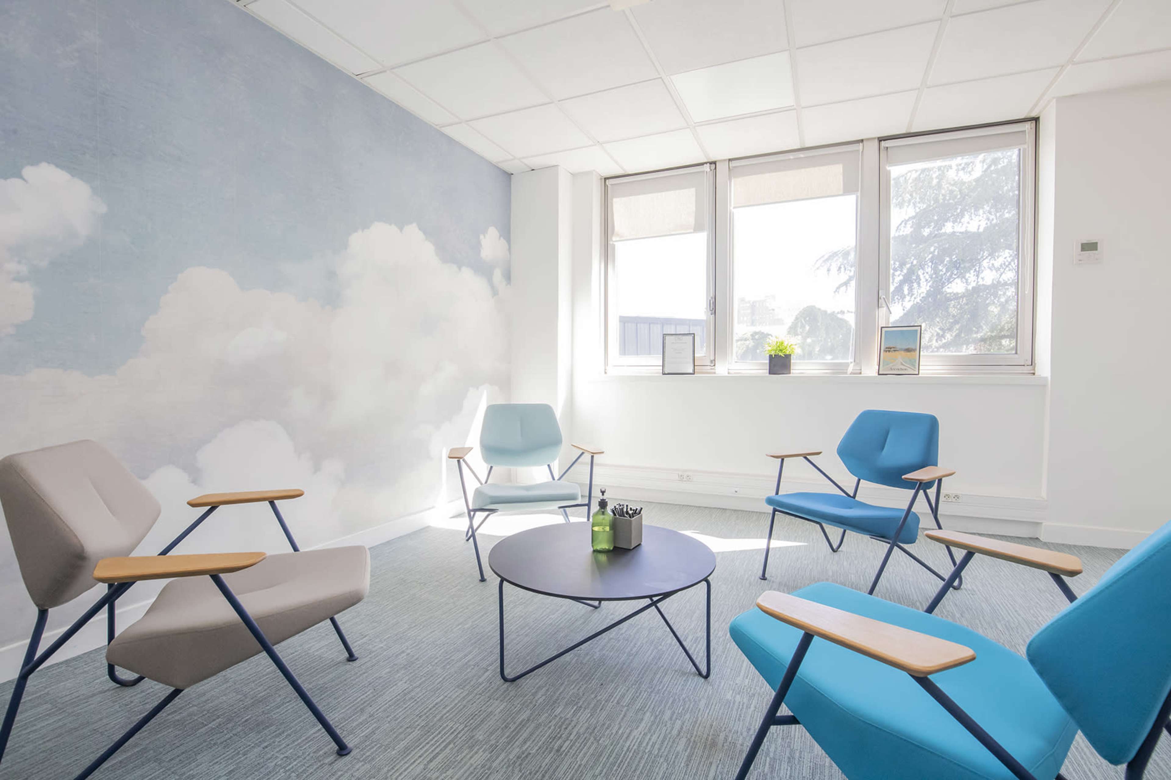 A light-filled room with four chairs arranged around a black circular table, featuring a mural of clouds on one wall.