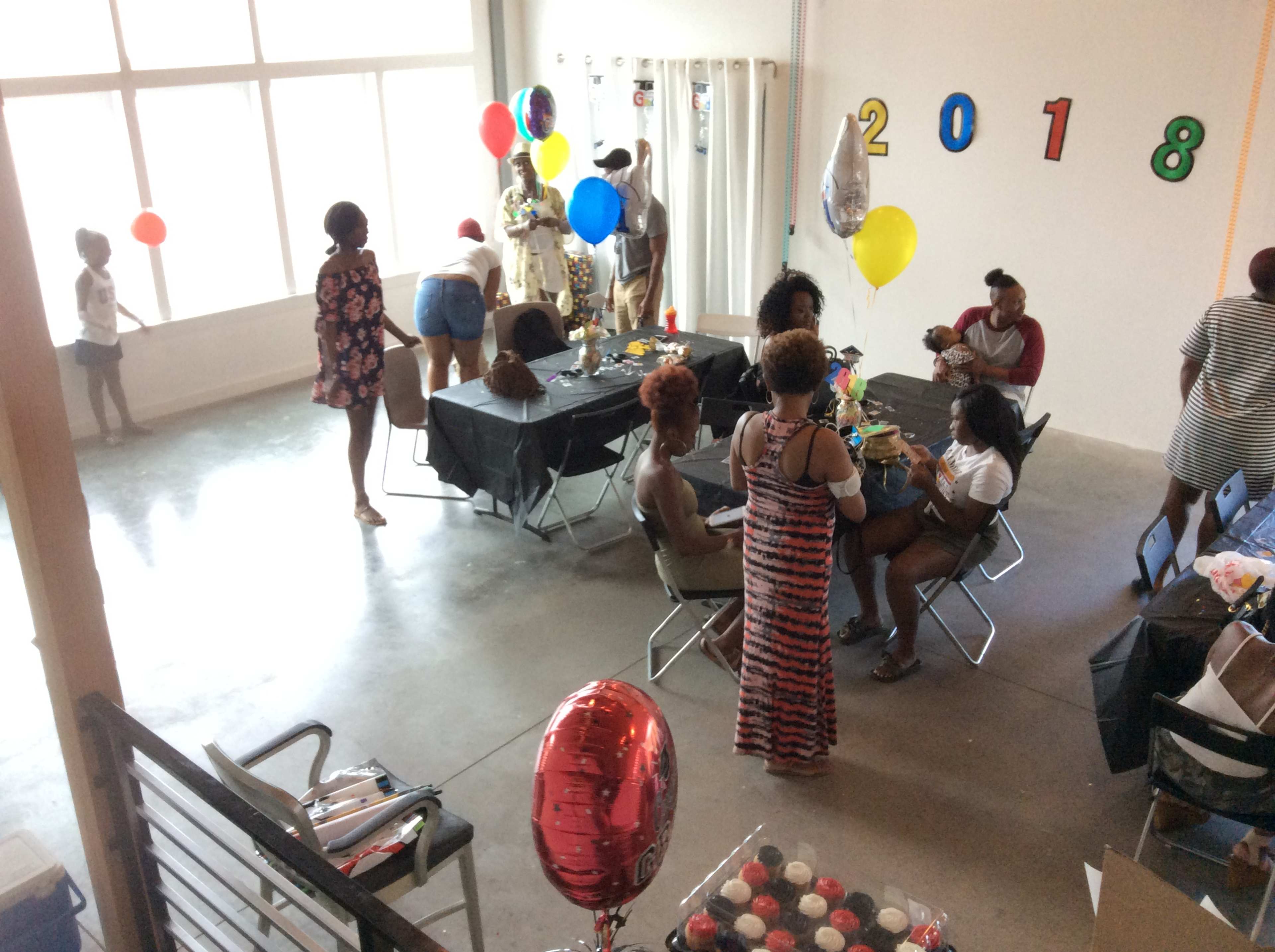 A group of people gathers in a spacious room decorated with balloons and a "2018" sign, engaged in a celebration around tables.