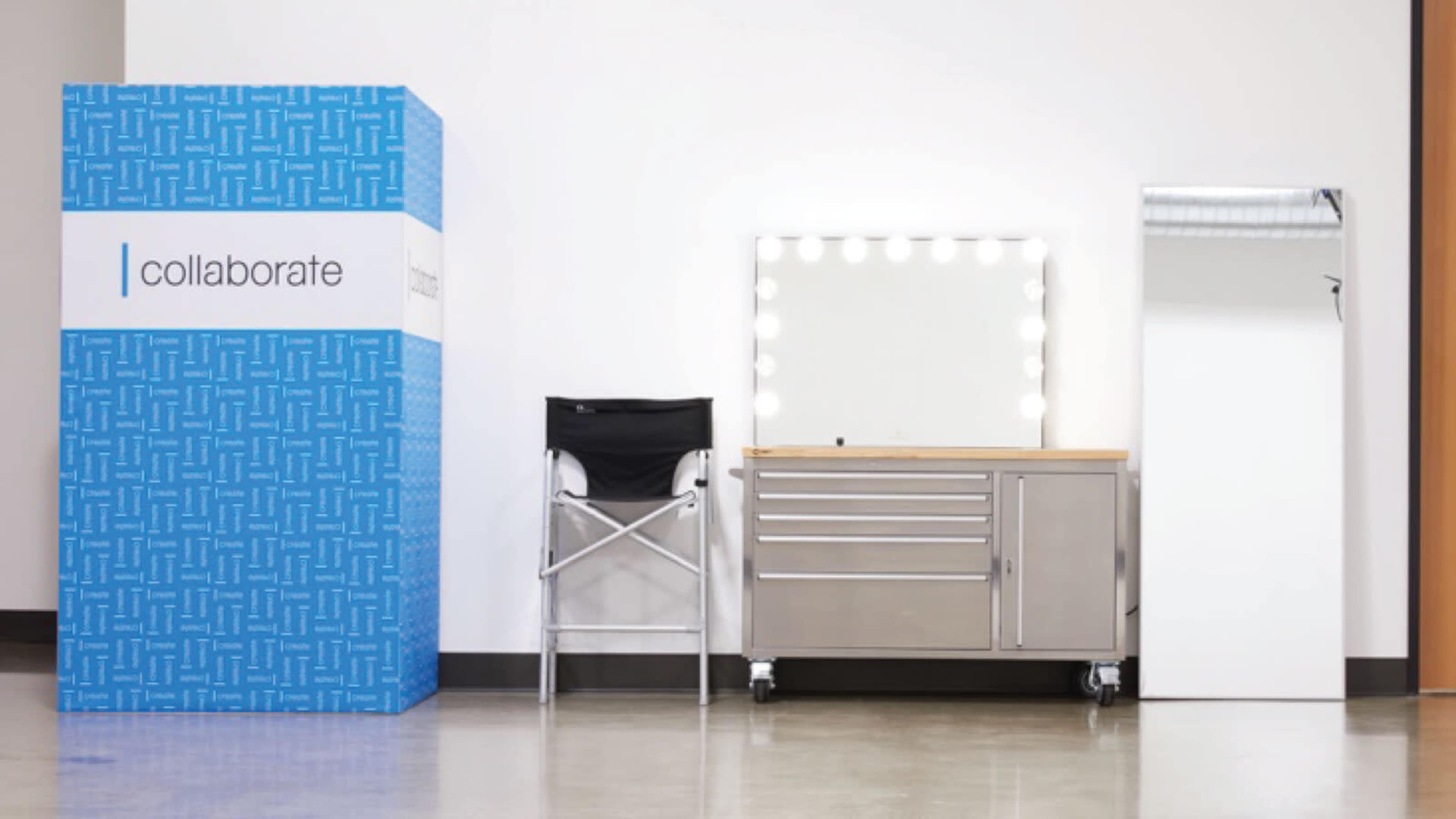 The image features a blue display box labeled "collaborate," a silver vanity with a mirror and lights, a rolling cart, and a tall mirror positioned against a wall.