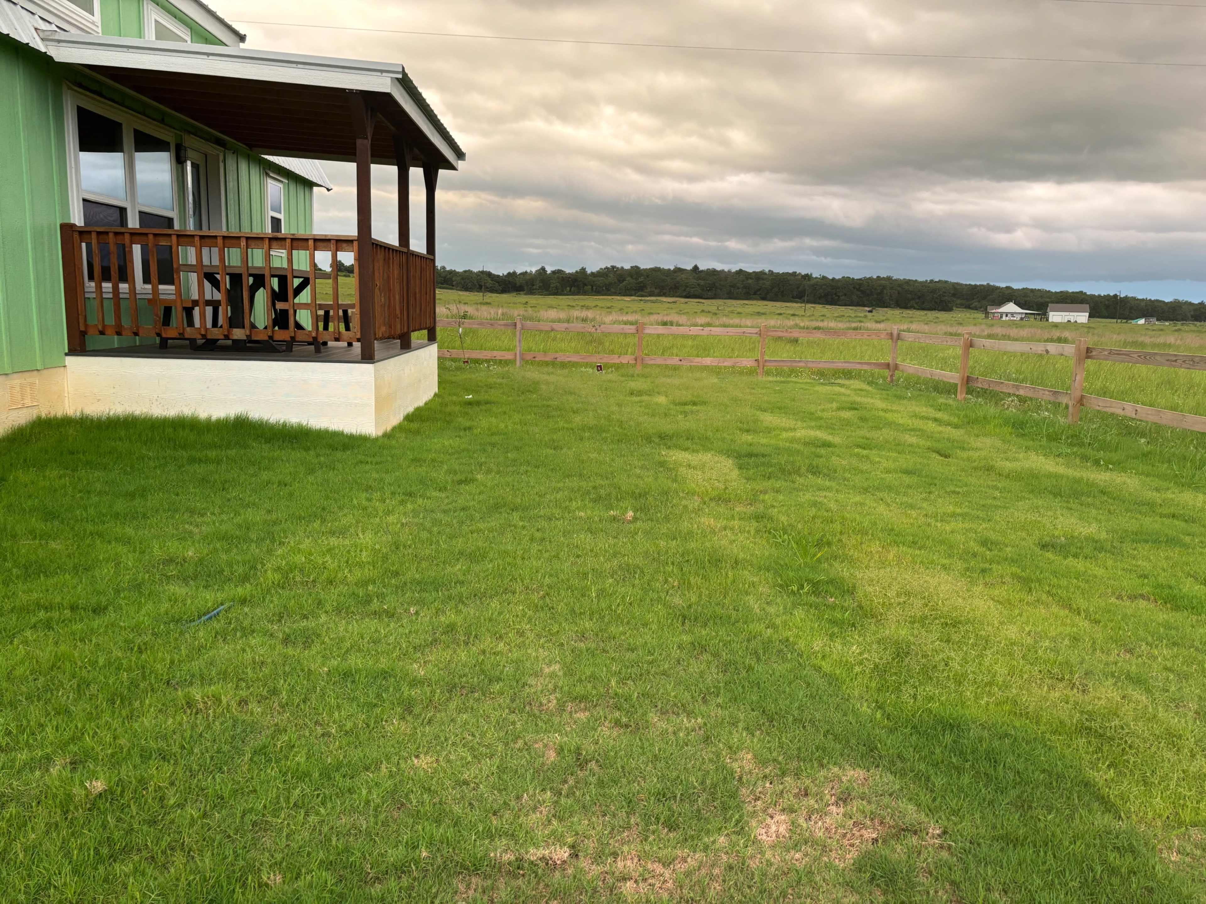 A green lawn stretches in front of a wooden porch attached to a light green house, with a wooden fence in the background against a cloudy sky.