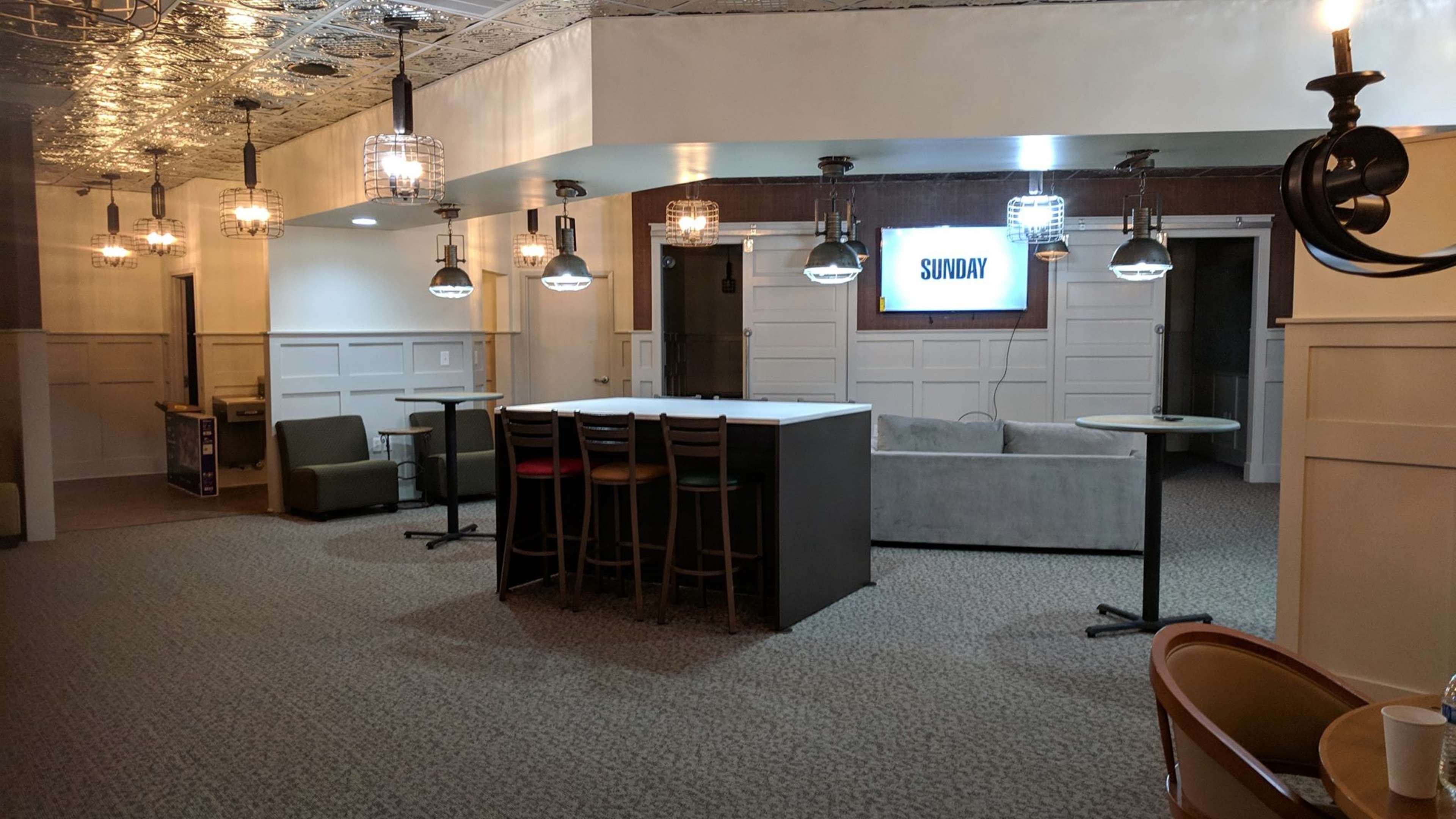 A spacious, modern lounge area with a central bar, high tables, and a screen displaying "SUNDAY" on one wall, illuminated by pendant lights.