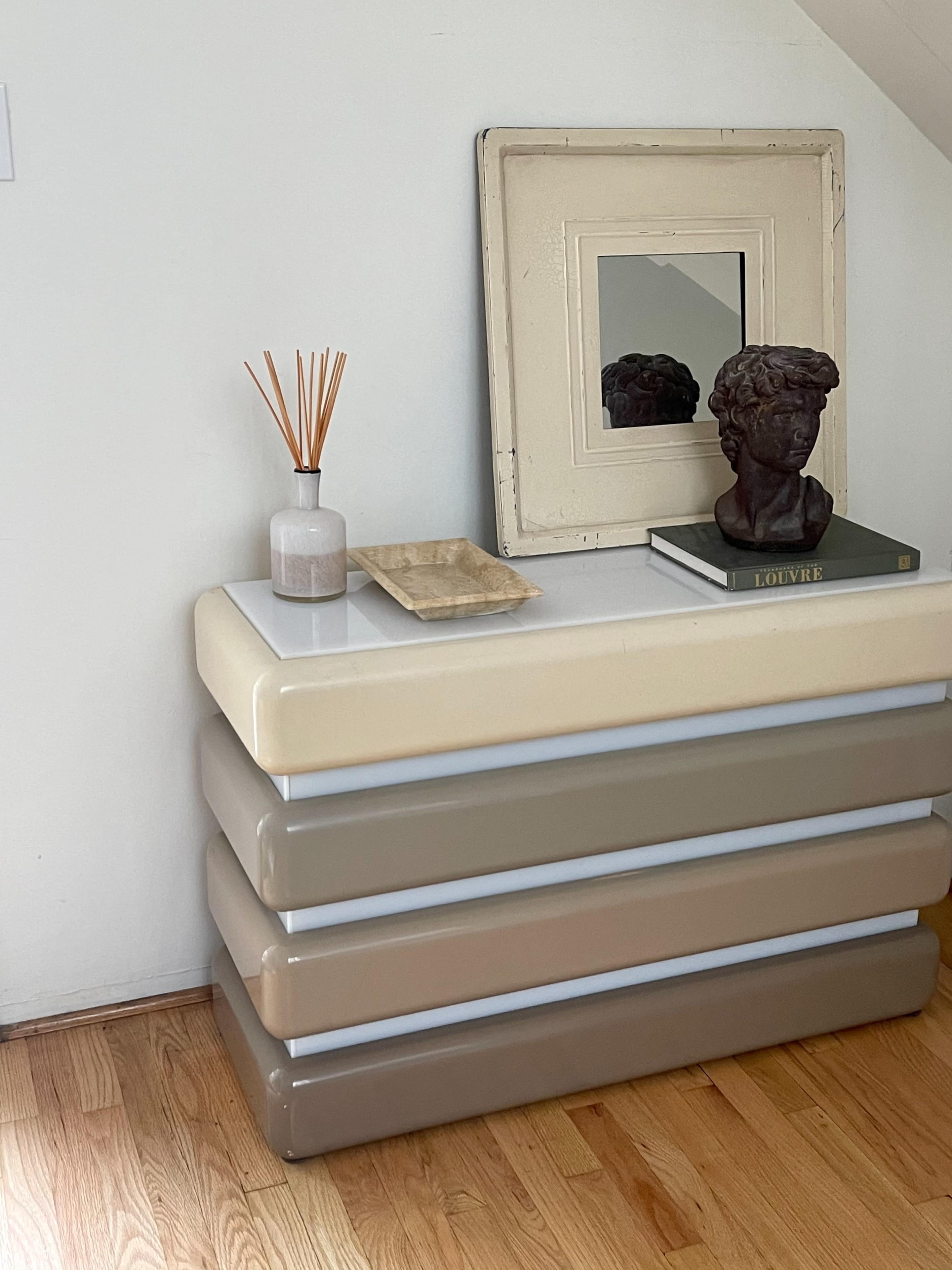 A modern two-tone console table with rounded layers sits beside a framed mirror and a decorative bust on a wooden floor.