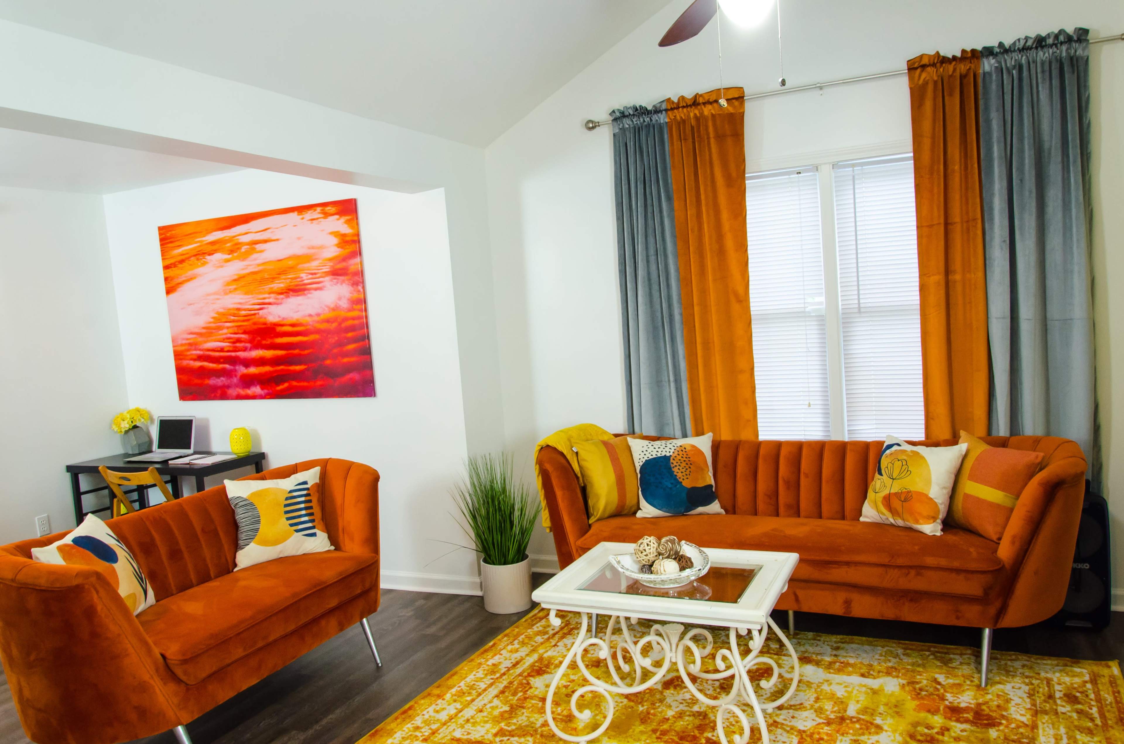 A living room features orange velvet sofas with decorative pillows, a glass coffee table, and colorful artwork on the wall, complemented by large windows with light-filtering curtains.