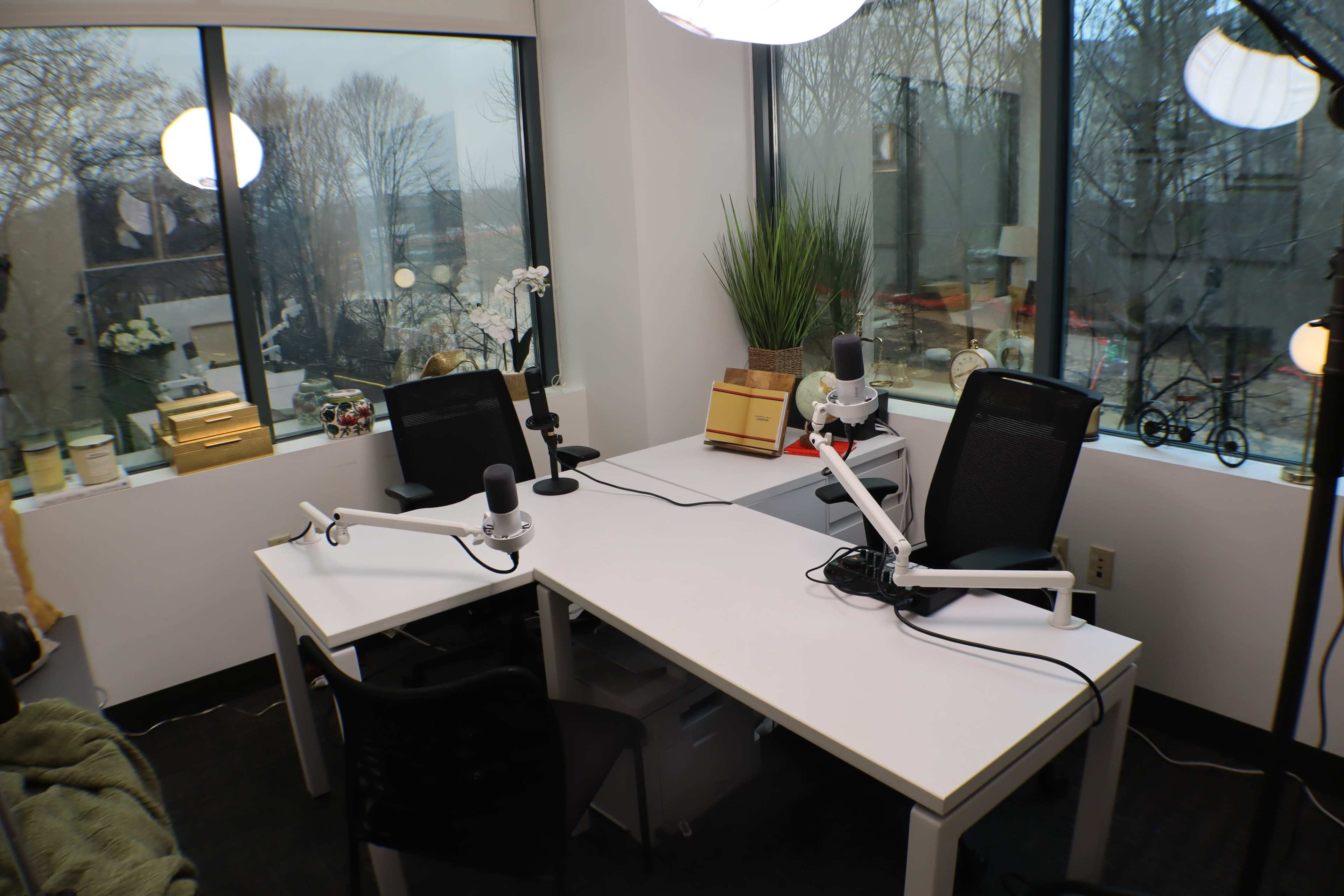 The image shows a modern podcasting studio featuring two desks with microphones, chairs, and large windows overlooking a wooded area.