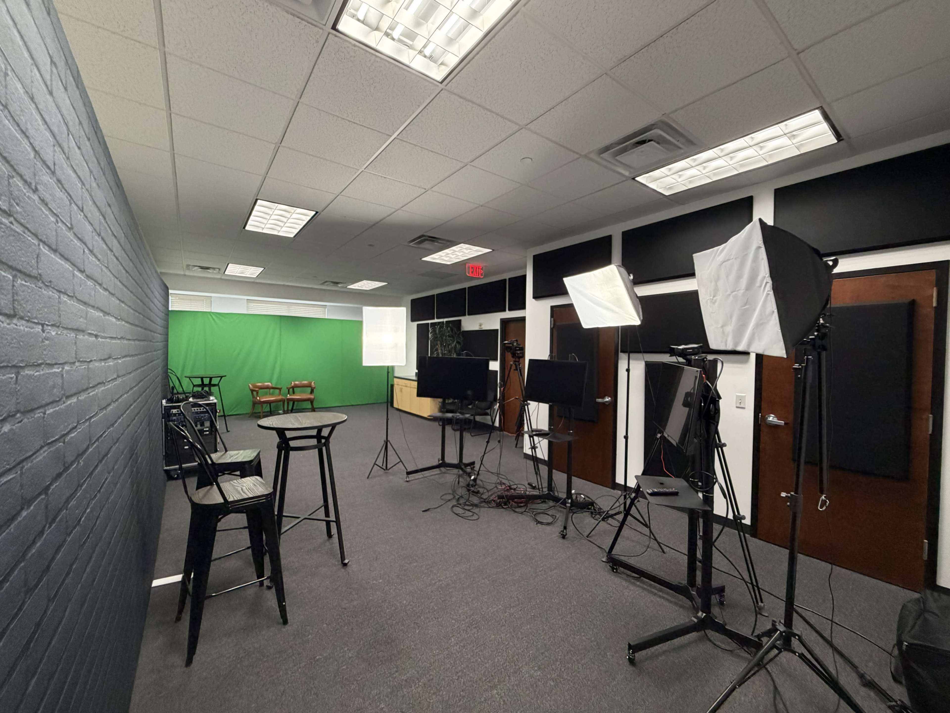 The image shows a studio setting with a green screen, cameras on tripods, lighting equipment, and a couple of chairs arranged in the background.