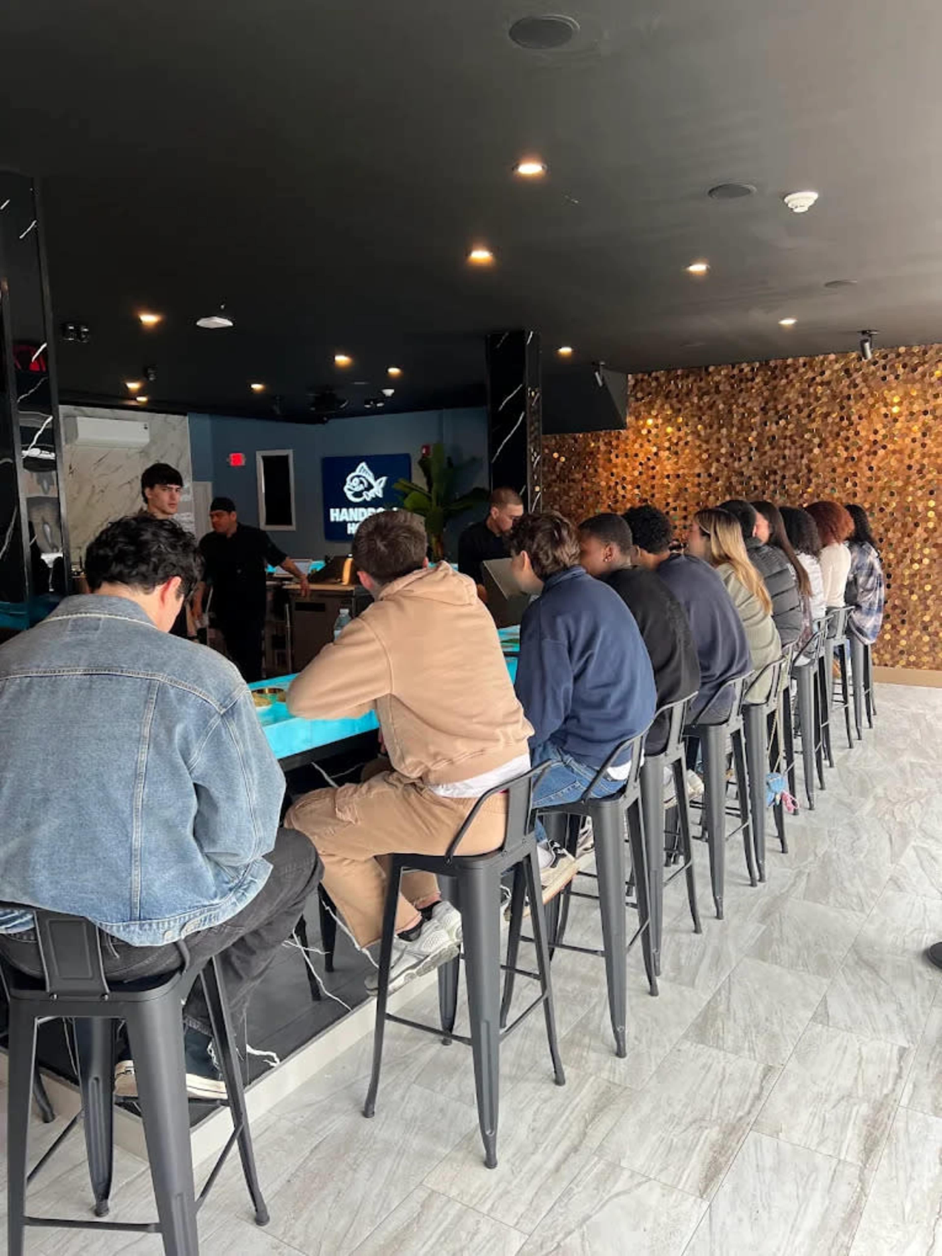 A group of young adults sits at a long bar counter in a modern interior, facing a screen while a bartender prepares drinks.