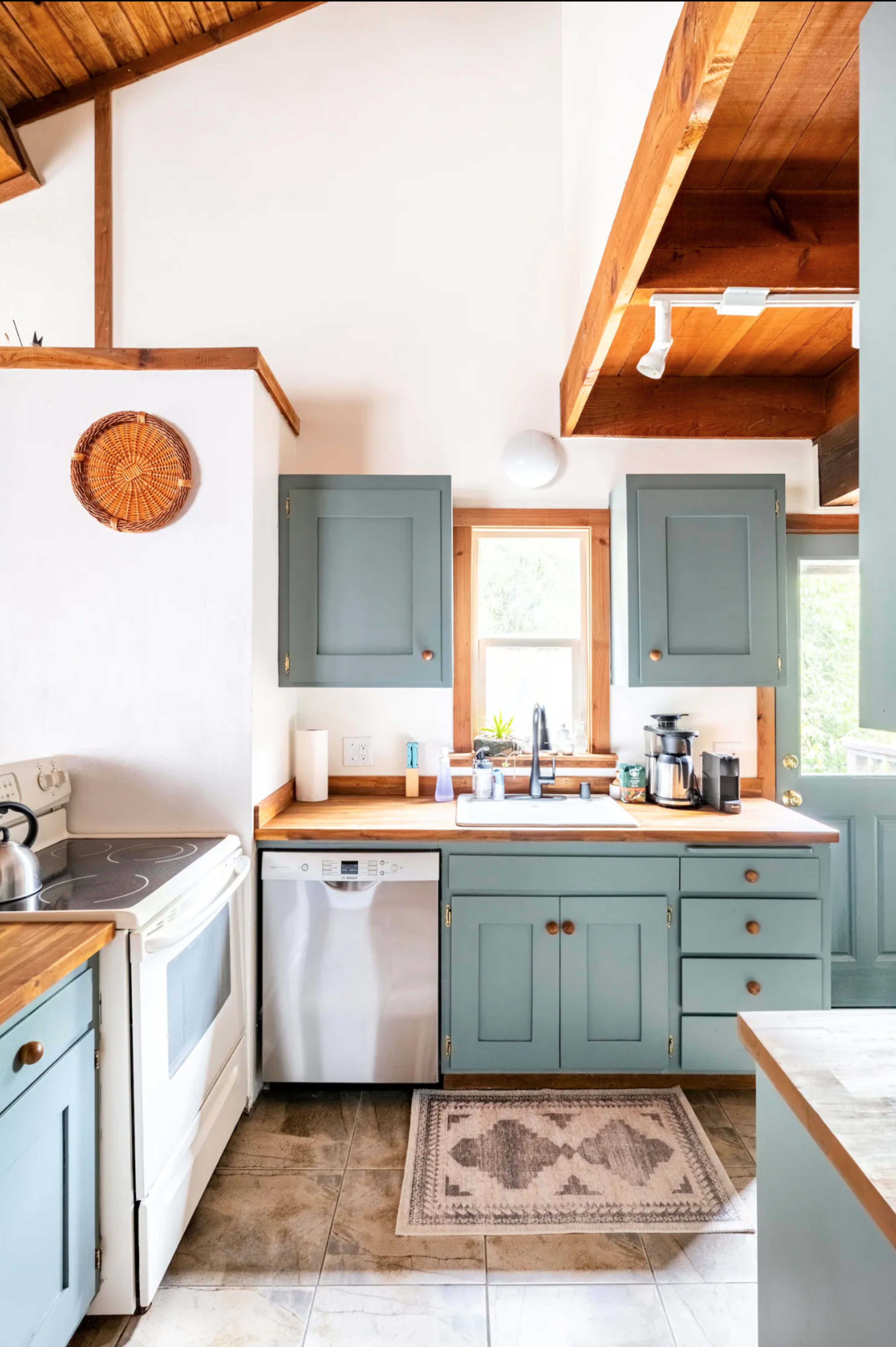 The image shows a kitchen with pale blue cabinets, a white stove, a dishwasher, and a wooden countertop, featuring natural light from a nearby window.