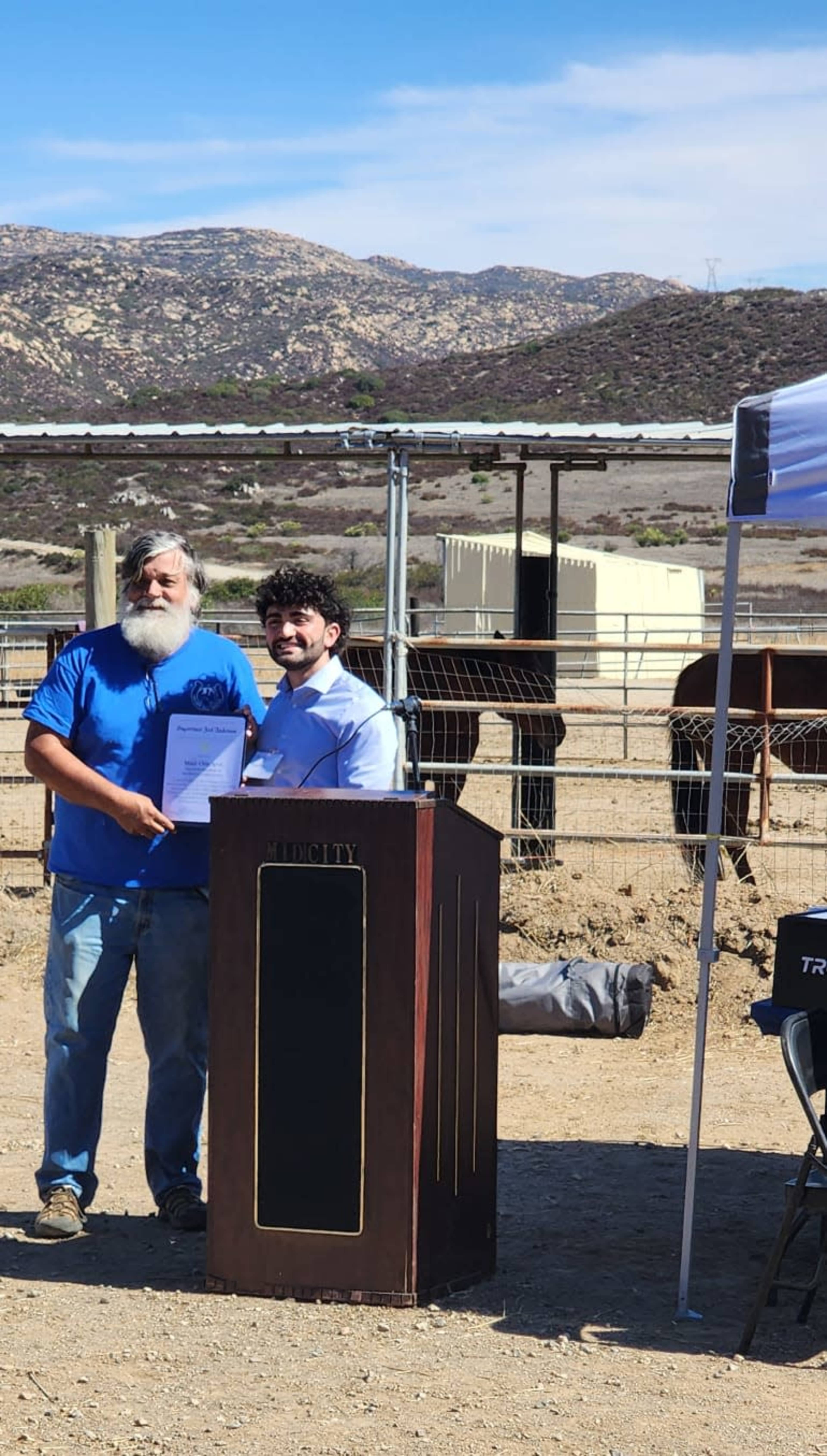 Two men stand together, one holding a document, in front of a podium, with horses and a mountainous landscape in the background.