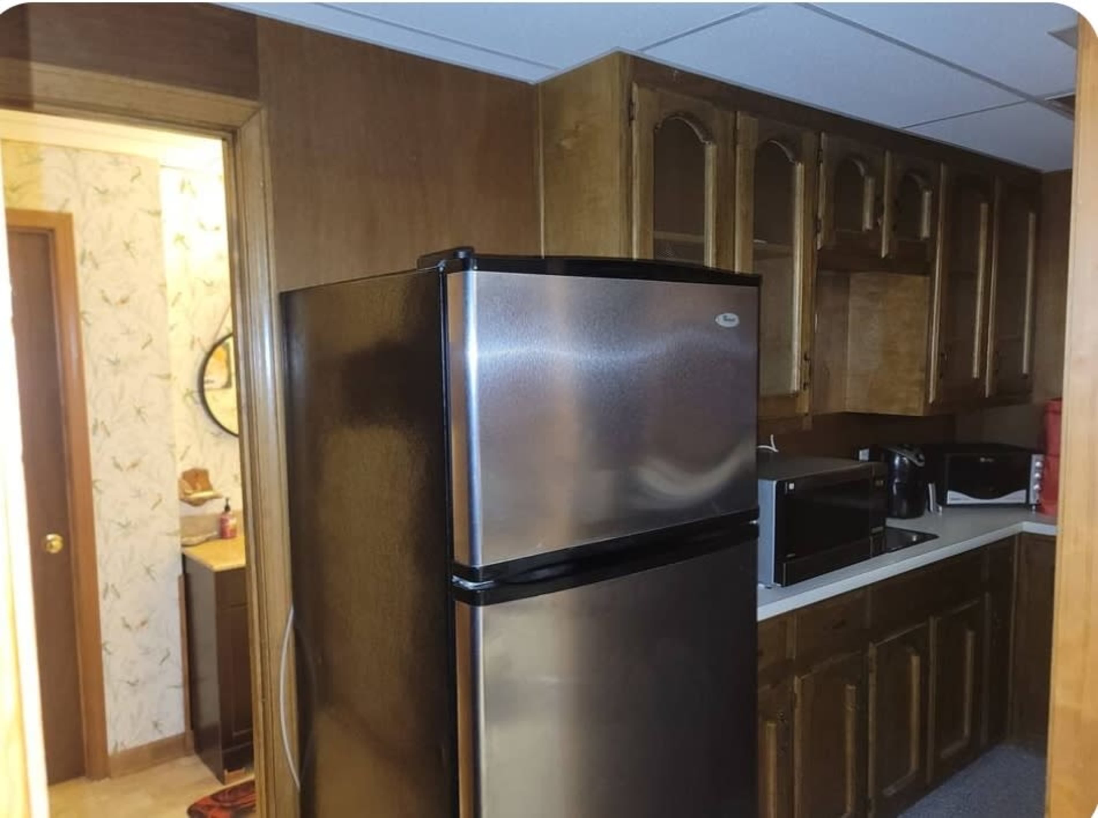 The image shows a kitchen area featuring a stainless steel refrigerator, a countertop with various appliances, and a doorway leading to a bathroom.