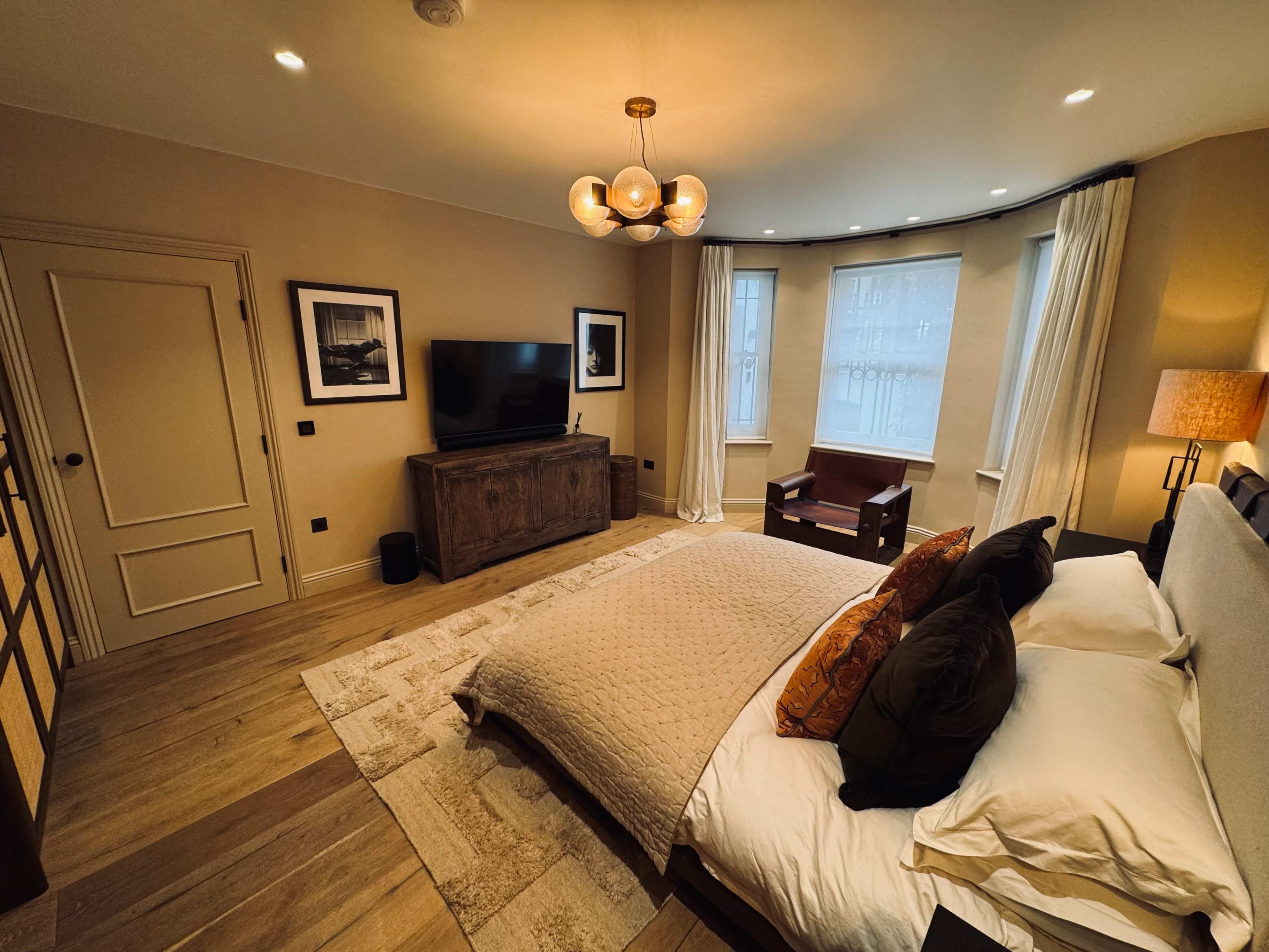 The image shows a stylish bedroom featuring a bed with decorative pillows, a wooden dresser, a television mounted on the wall, and a seated area by a large window.