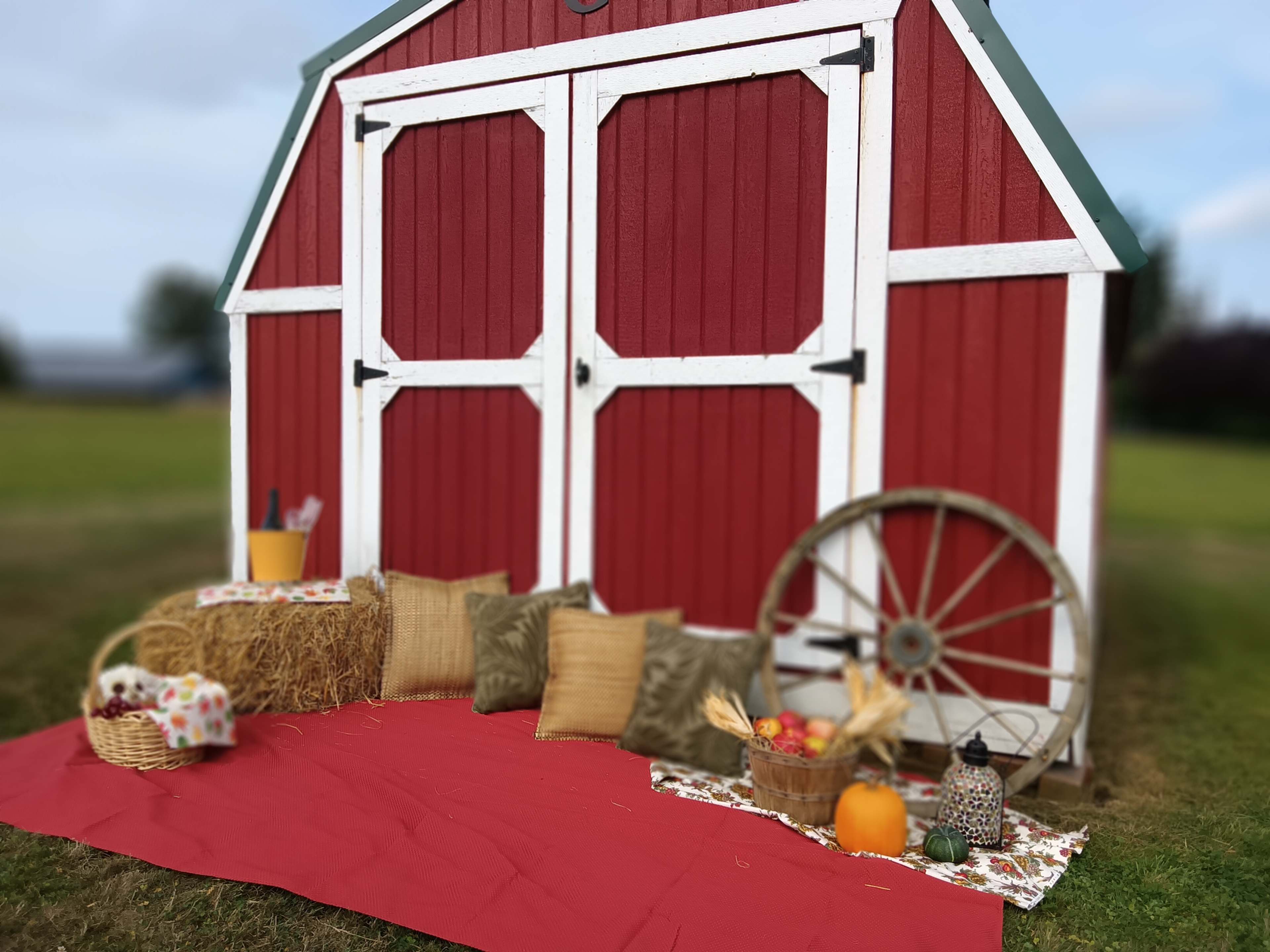 A red barn with white trim serves as a backdrop for a cozy outdoor seating area featuring hay bales, cushions, a decorative wheel, and autumn-themed decorations.