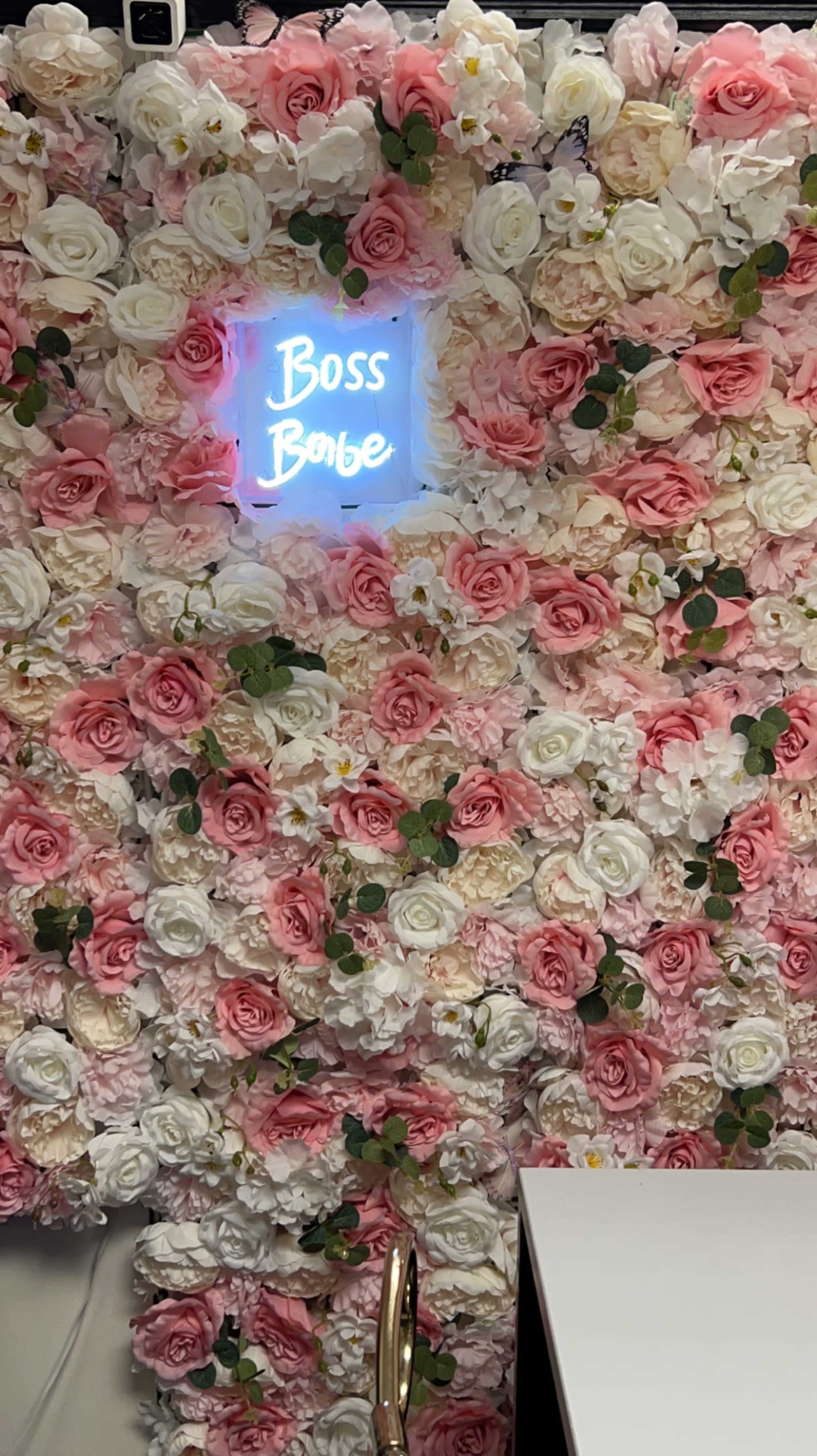 The image features a wall covered in artificial flowers, primarily in shades of pink and white, with a neon sign that reads "Boss Babe."