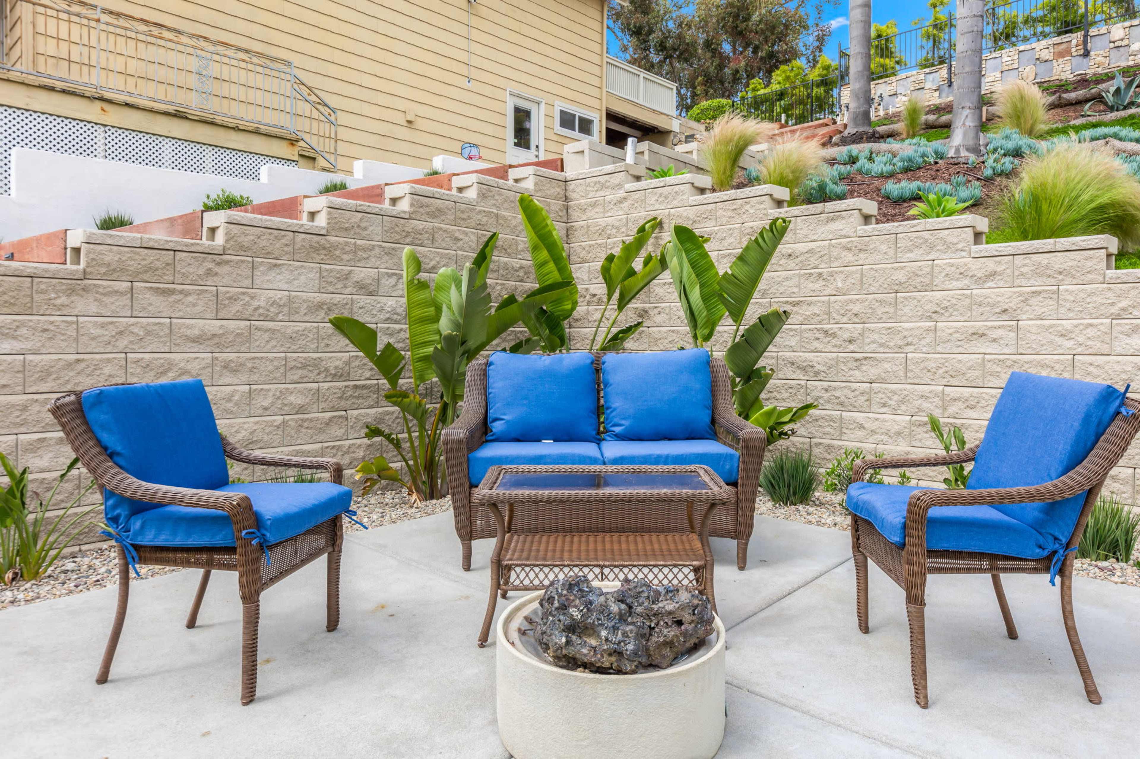A patio area features a set of blue cushions on wicker furniture arranged around a glass-top coffee table, with plants and landscaping in the background.