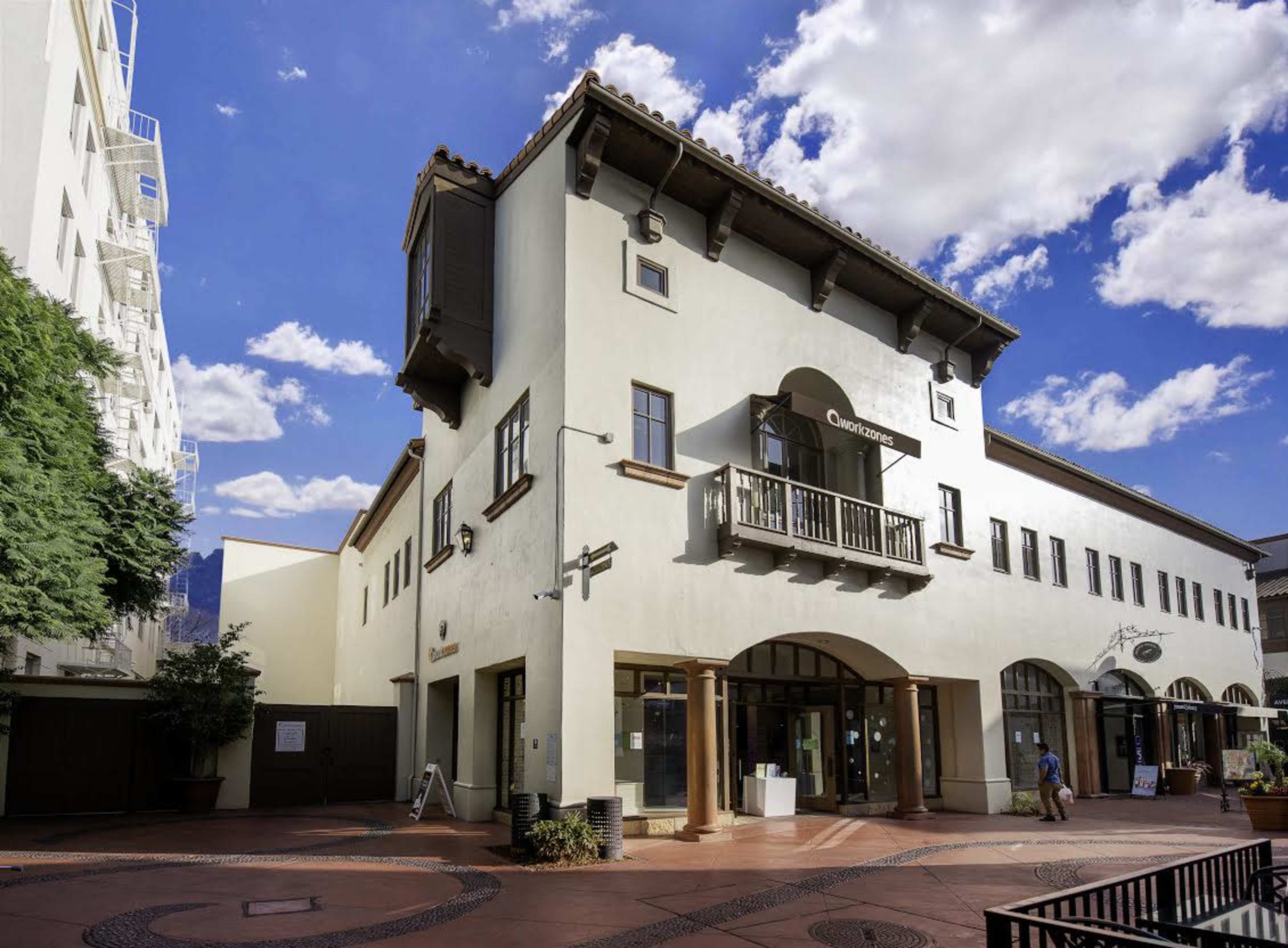 A three-story building with a balcony and arched entrance, situated in an outdoor plaza under a partly cloudy sky.
