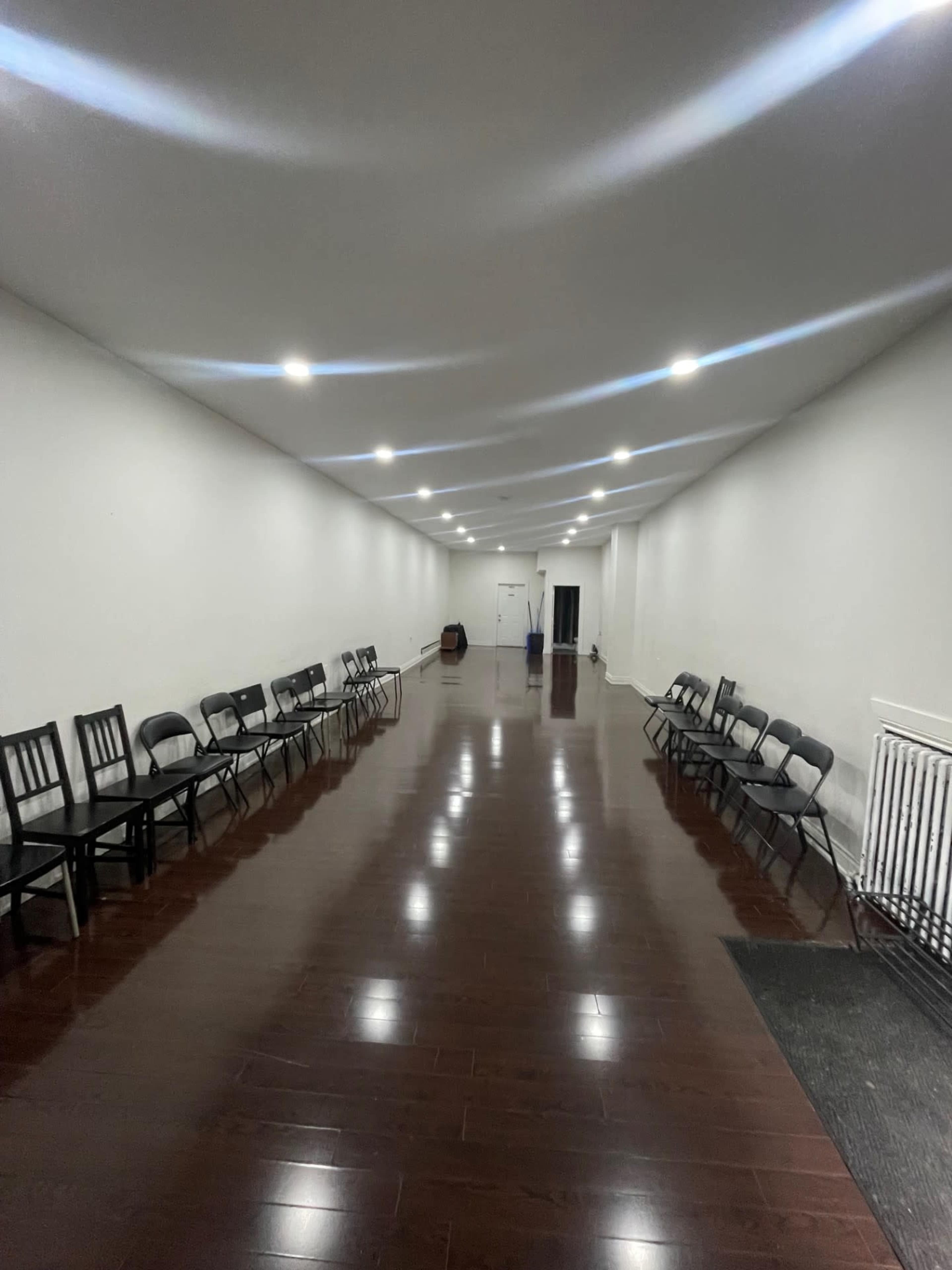 The image shows a long, narrow room with glossy wooden flooring lined with black chairs on either side and bright overhead lights illuminating the space.
