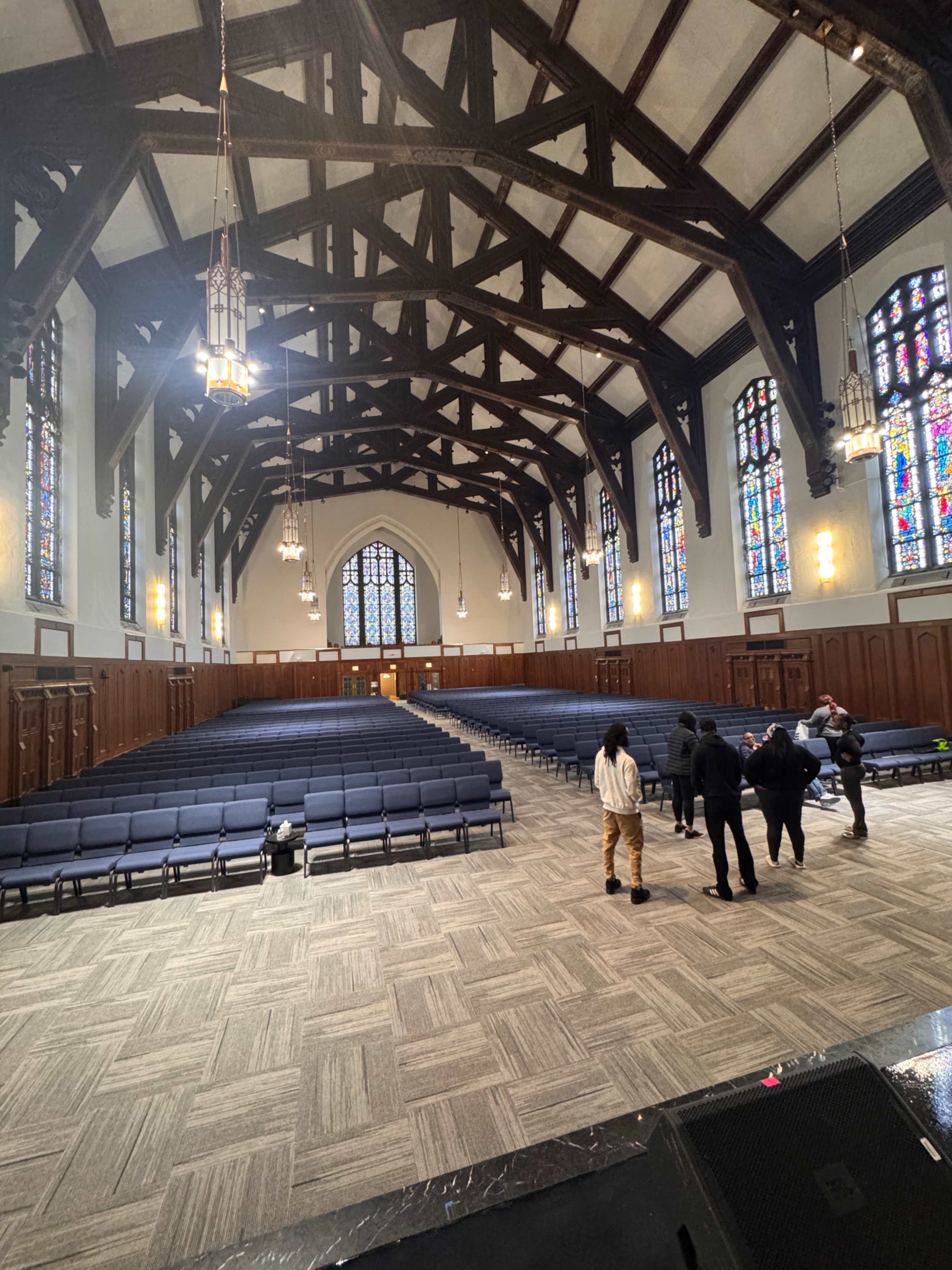 The image shows the interior of a large, empty hall featuring high wooden beams, stained glass windows, and rows of seating.