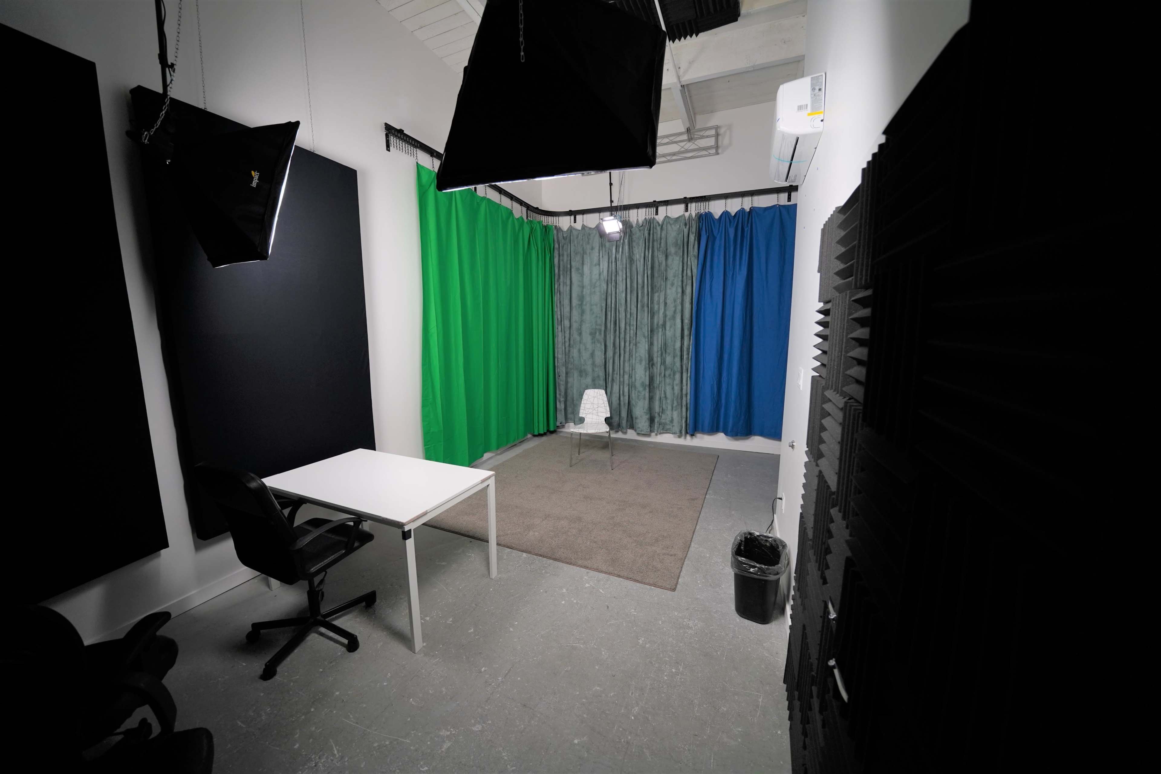 A video production studio featuring a white table and chair on a carpet, surrounded by green, blue, and gray curtains, with softbox lighting elements above.