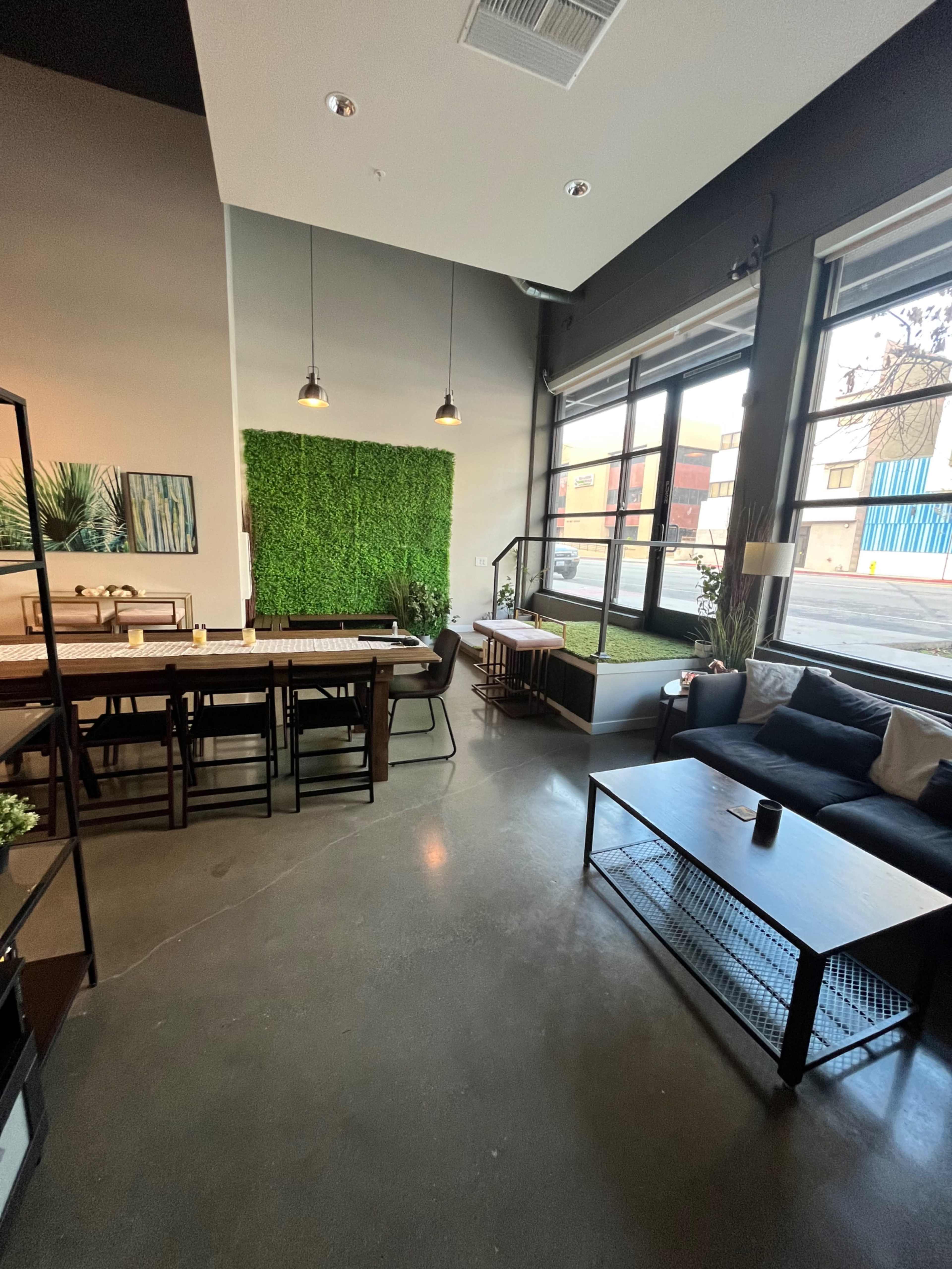A modern café interior with a green living wall, a long wooden table, and a cozy seating area near large windows.