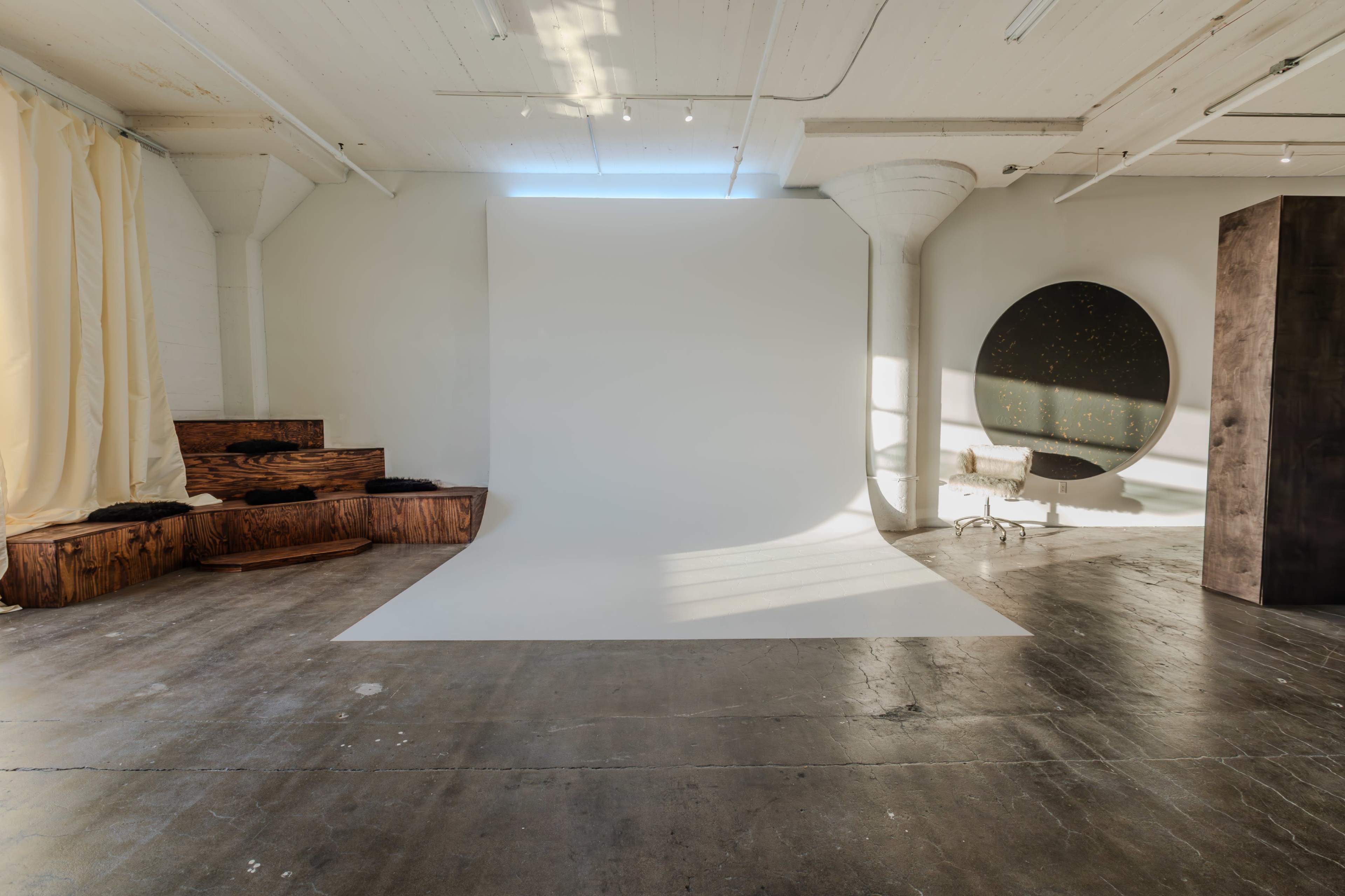 A spacious studio with a blank backdrop, wooden seating, and a circular mirror reflecting light.
