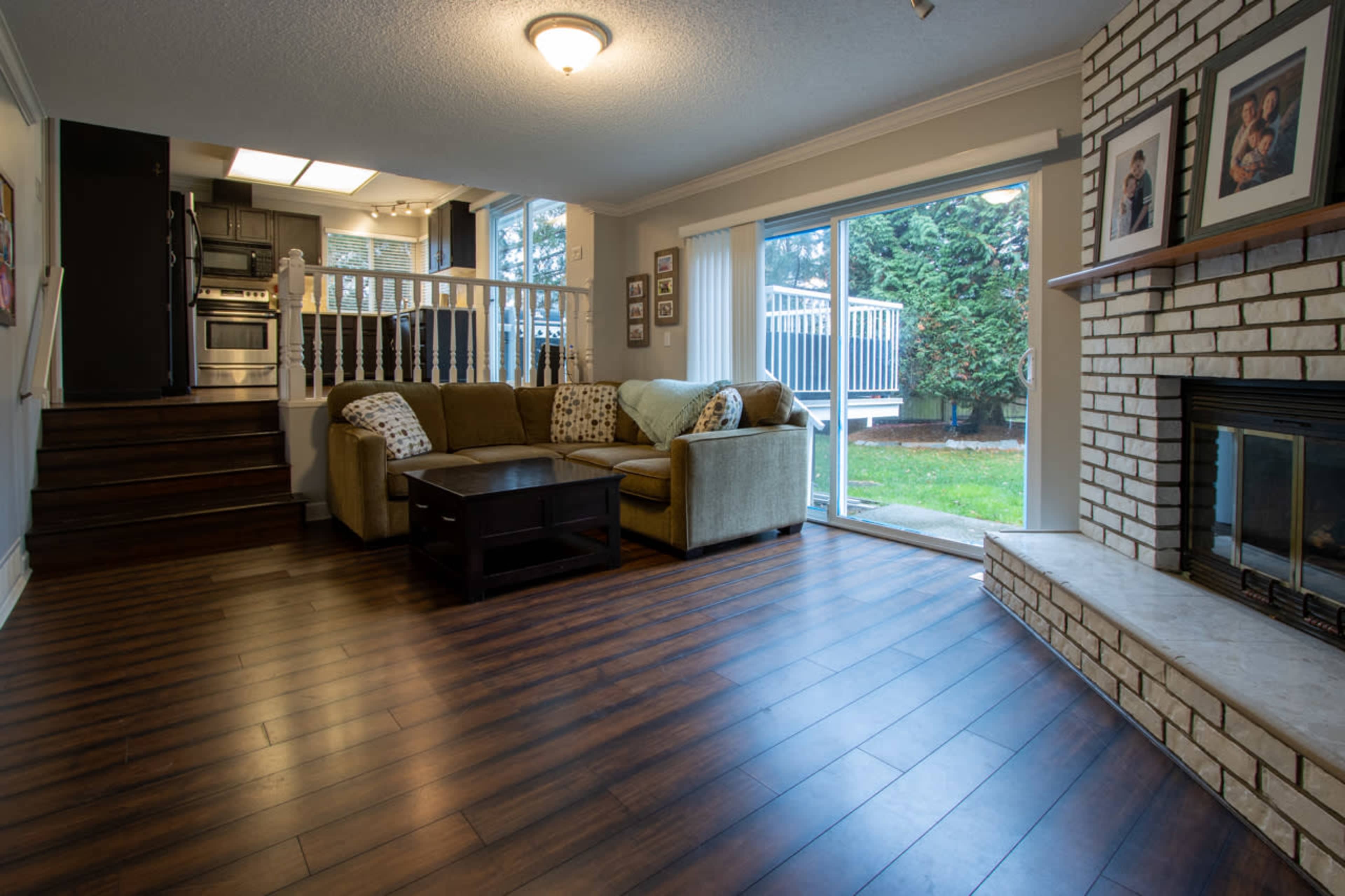 A cozy living room features a sofa, a coffee table, and a stone fireplace, with large windows overlooking a grassy outdoor area.
