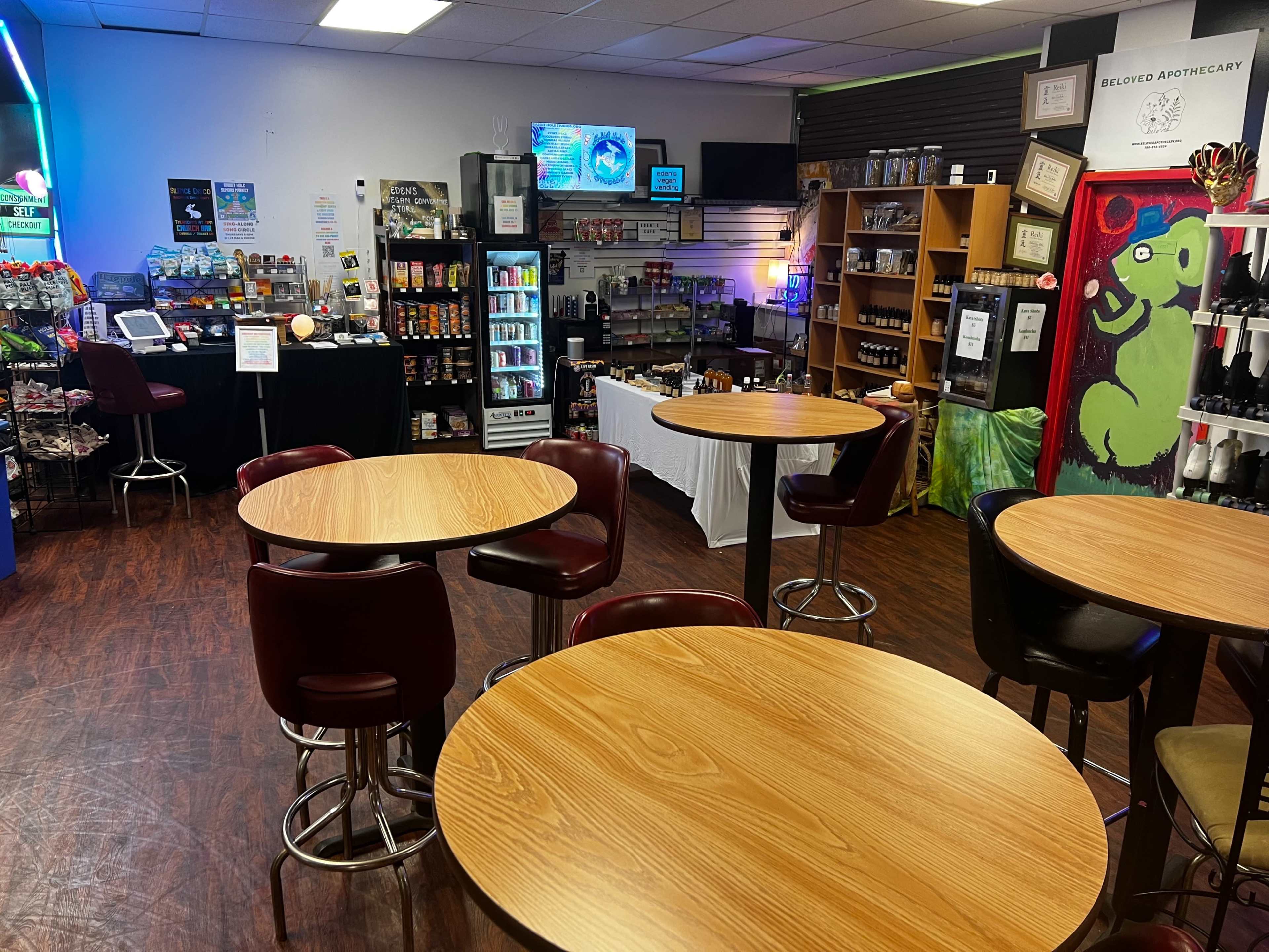The image shows a store interior with several wooden tables and chairs, display shelves filled with products, and a refrigerator in the background.