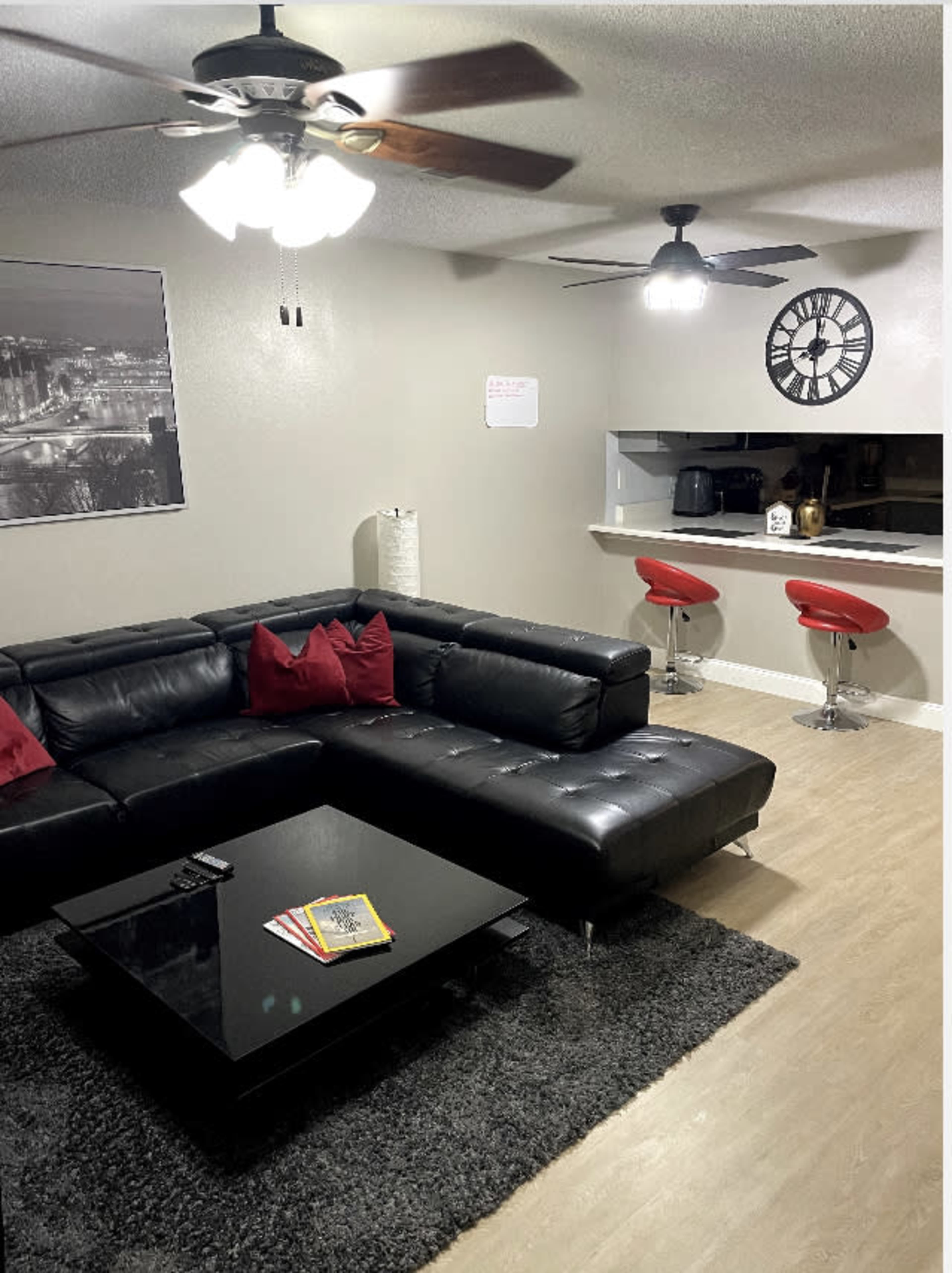 A modern living room features a black sectional sofa with red cushions, a glass coffee table on a rug, and a bar area with red stools in the background.