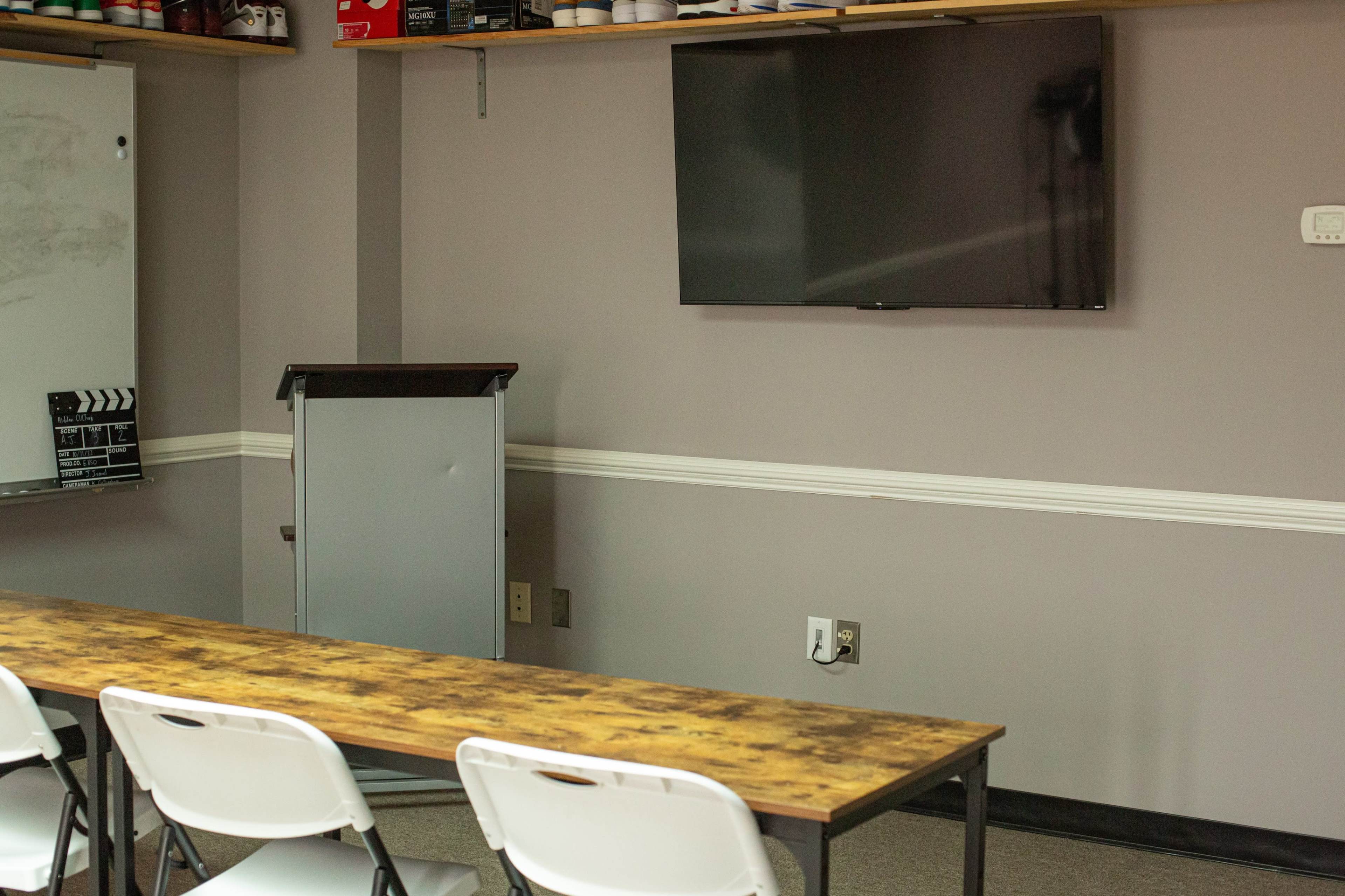 A classroom setting with a long table, white chairs, a television mounted on the wall, and a gray presentation board.