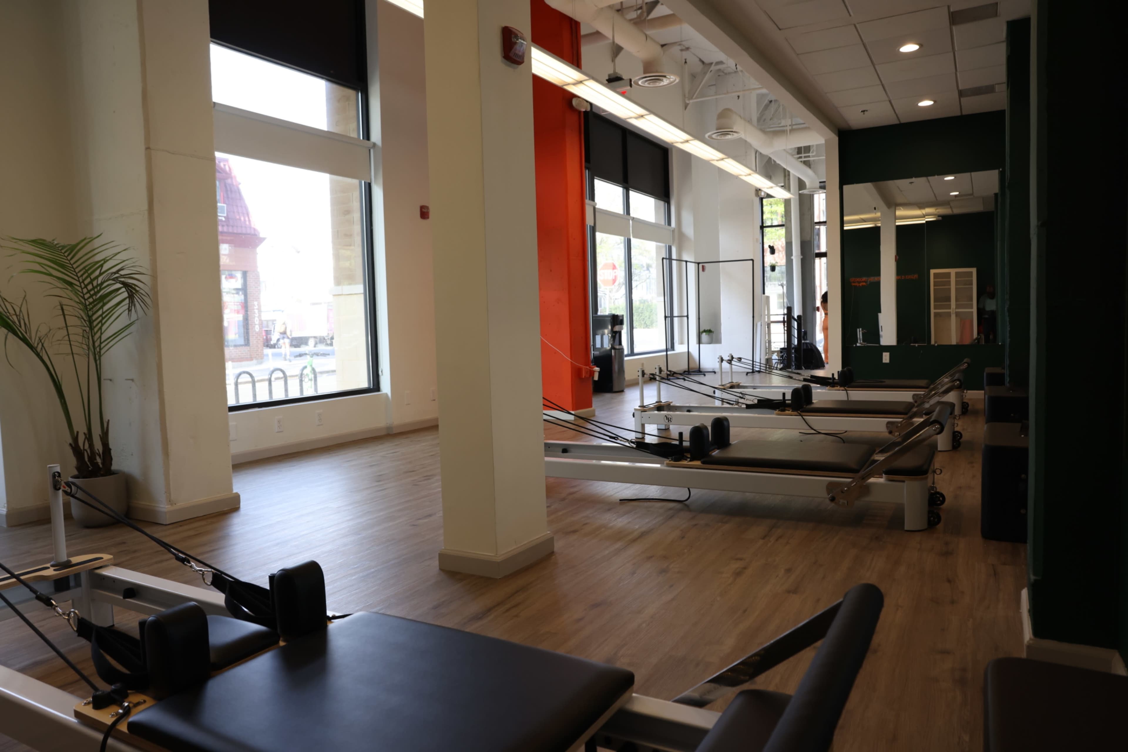 The image shows a spacious gym studio with reformers arranged neatly on a wooden floor and large windows allowing natural light to enter.