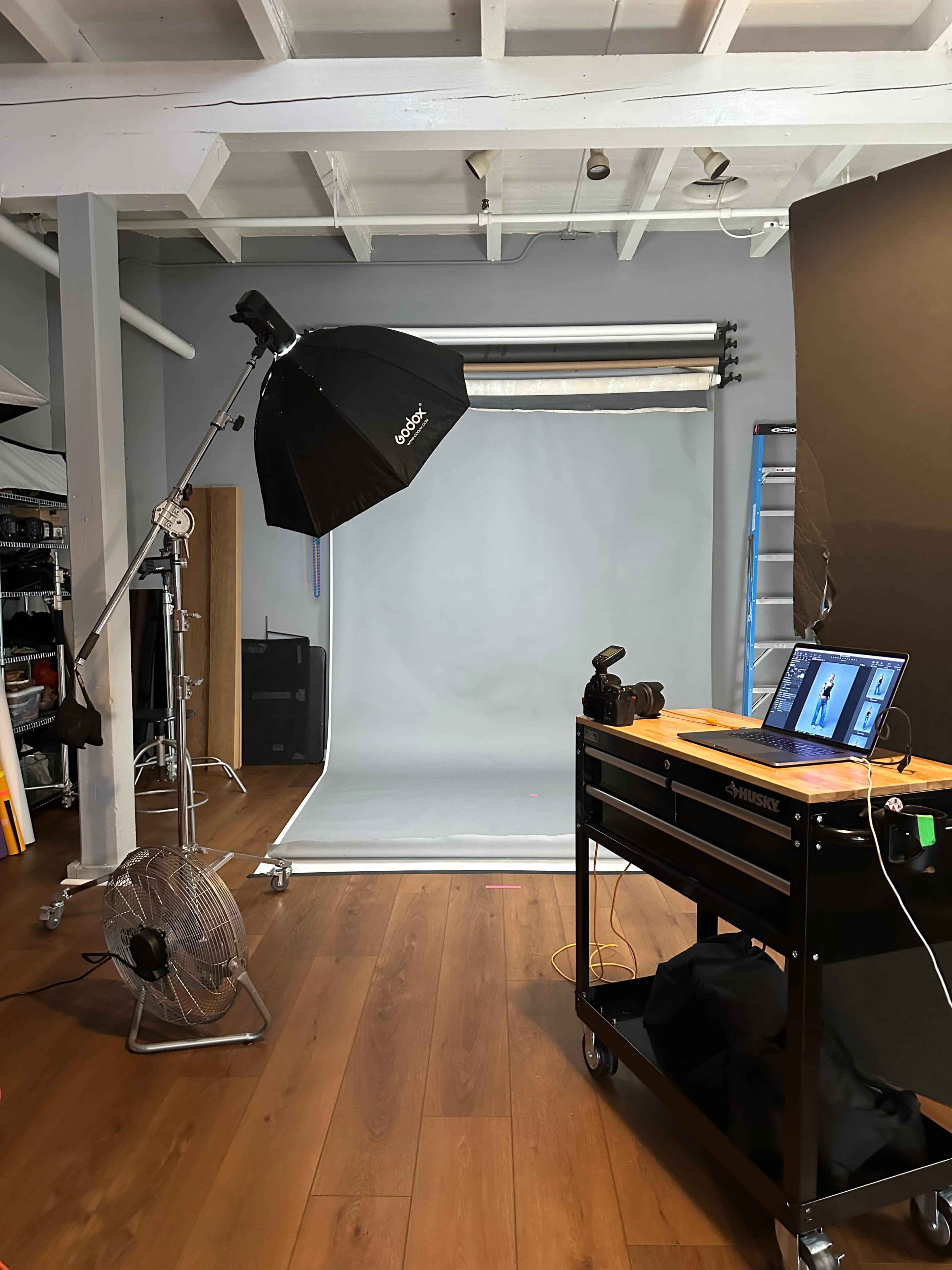 The image shows a photography studio equipped with a lighting umbrella, a backdrop, and a cart with a laptop and camera gear.