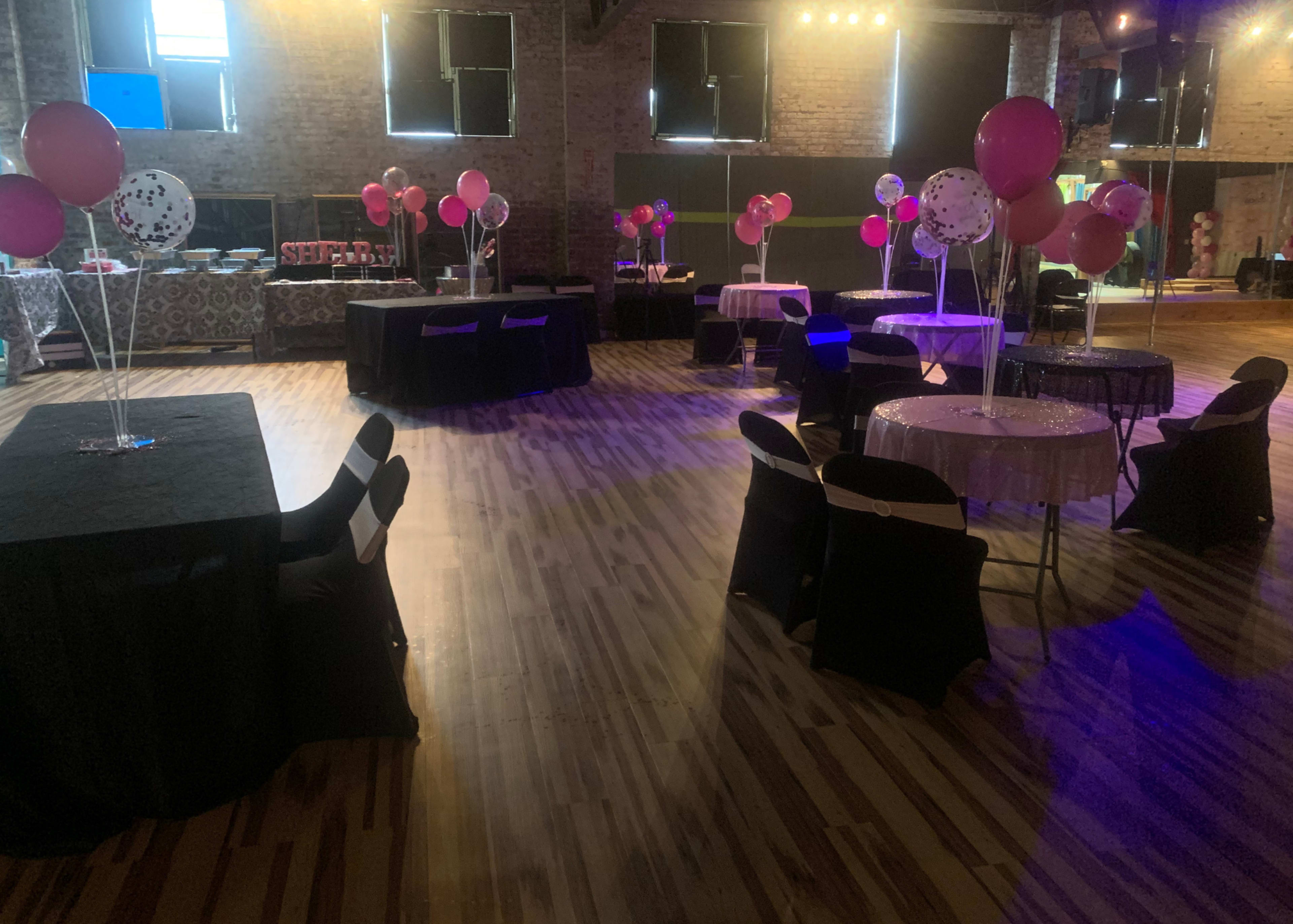 A spacious event venue features black and pink tables, balloons, and a polished wooden floor.