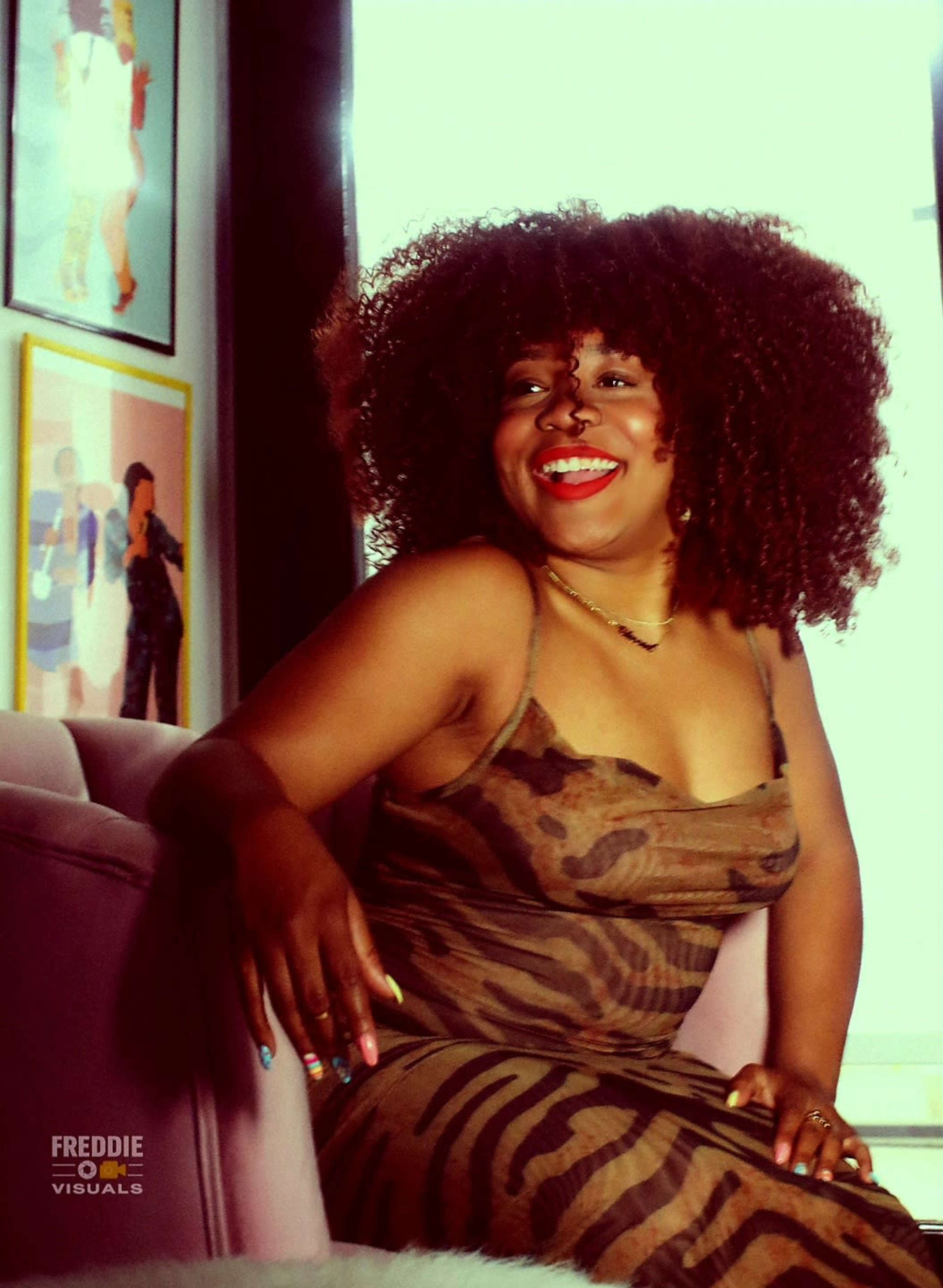 A person with curly hair sits on a couch, wearing a brown patterned dress, and smiles while posed against a backdrop of framed artwork.