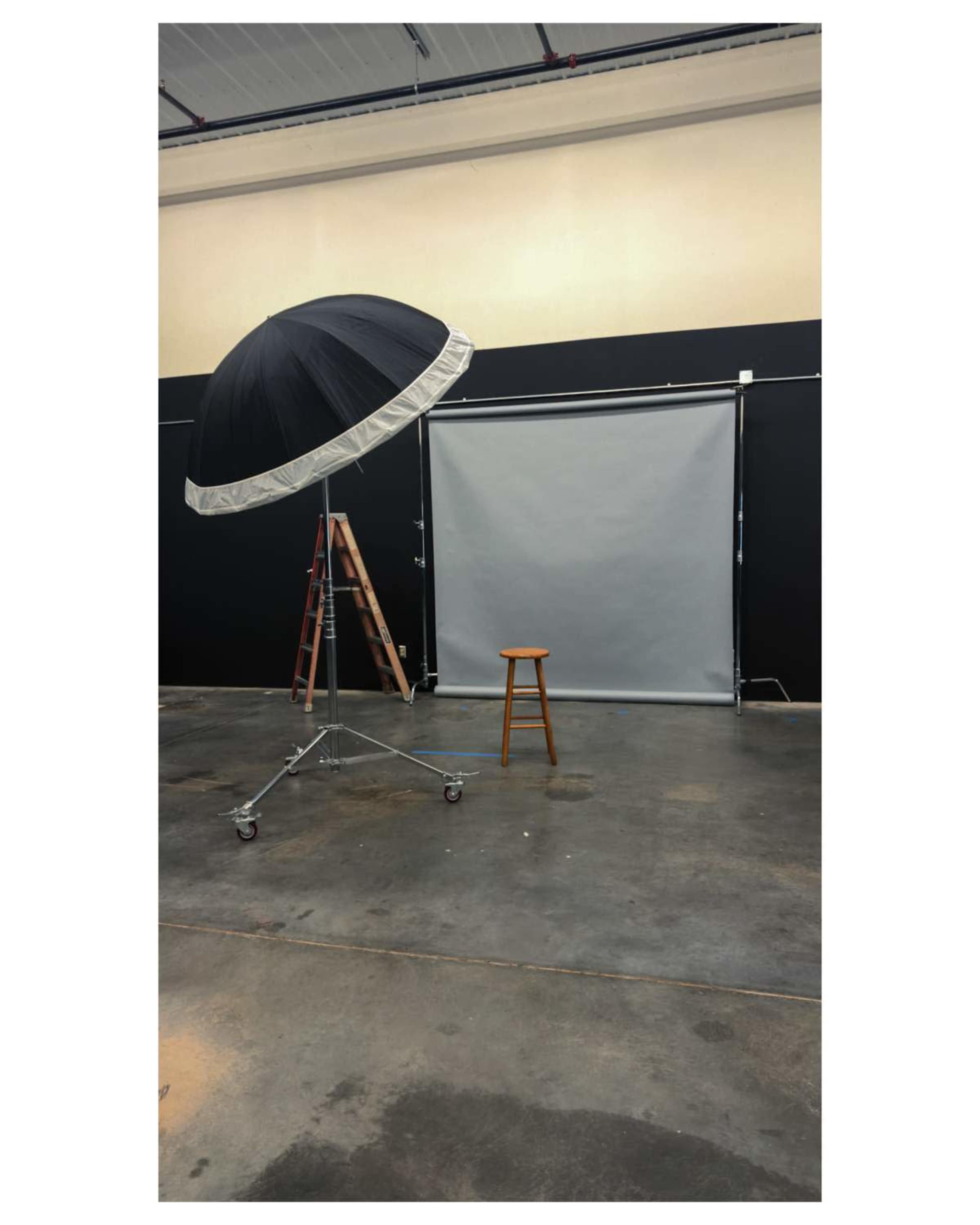 The image shows a photography studio with a large umbrella light, a gray backdrop, a wooden stool, and a ladder in the background.