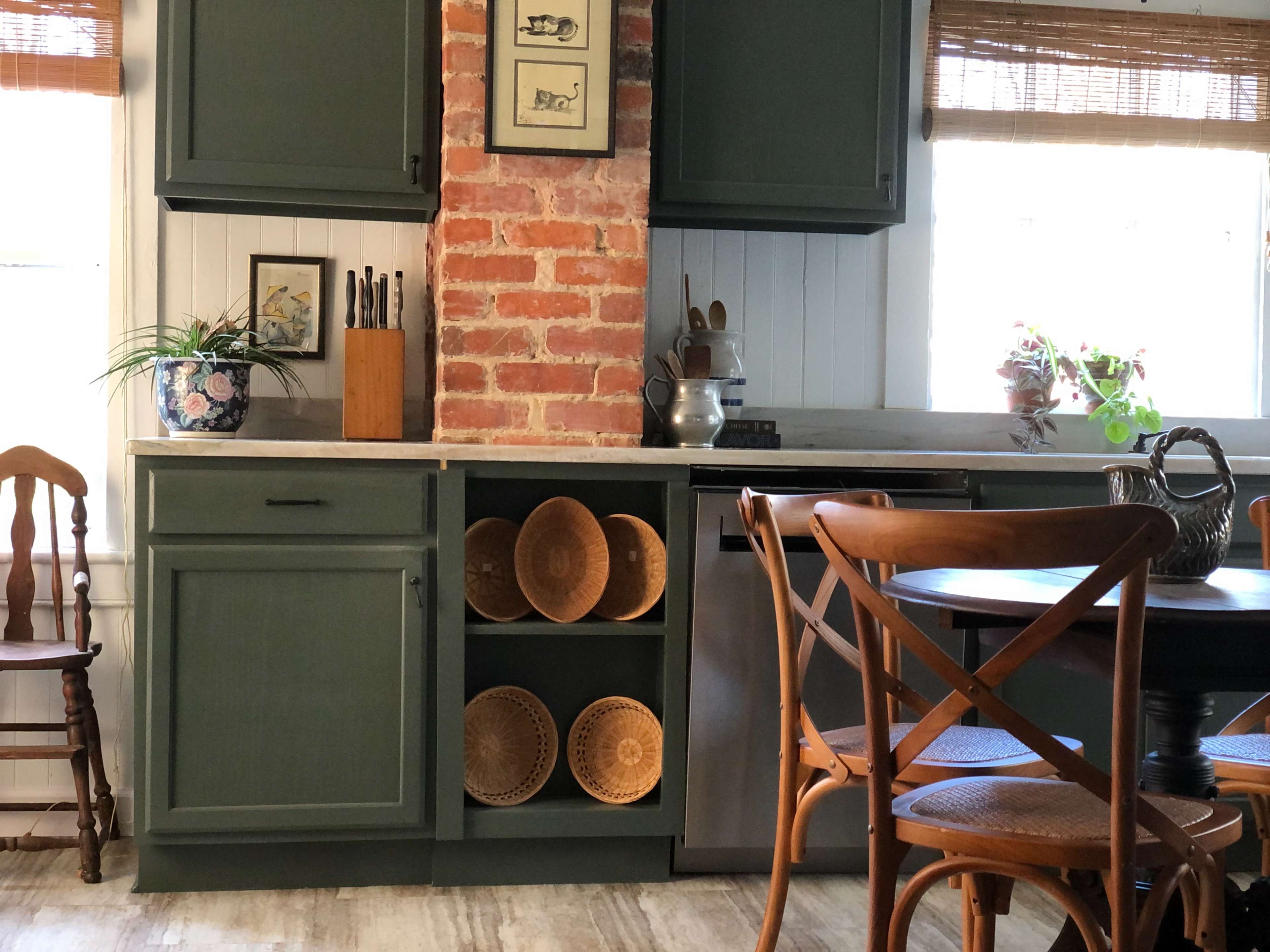 The image shows a kitchen with green cabinets, a brick column, wooden dining chairs, and decorative woven plates on display.