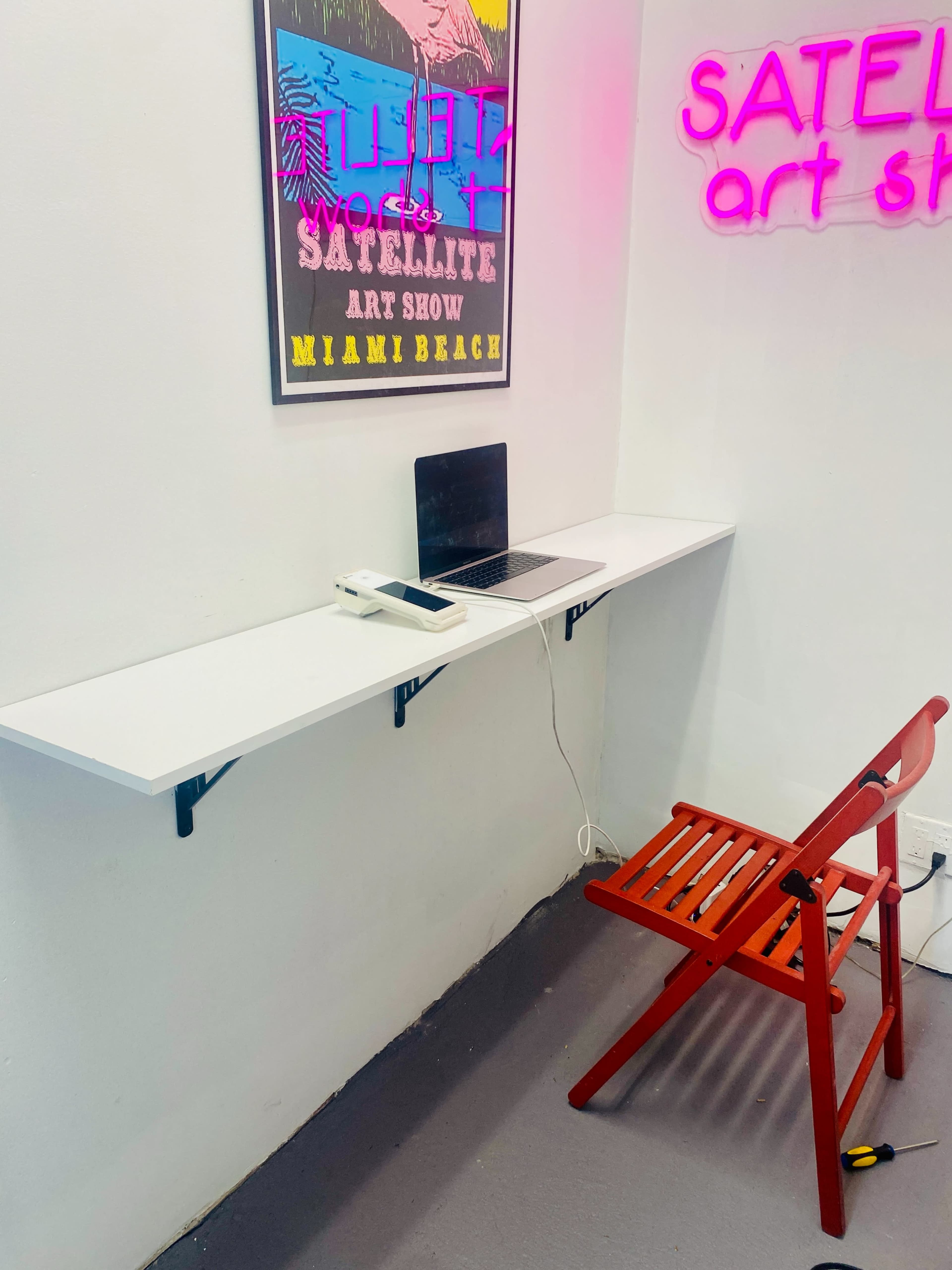 A white wall-mounted shelf with a laptop and charger above a red wooden chair in a room featuring a neon sign.