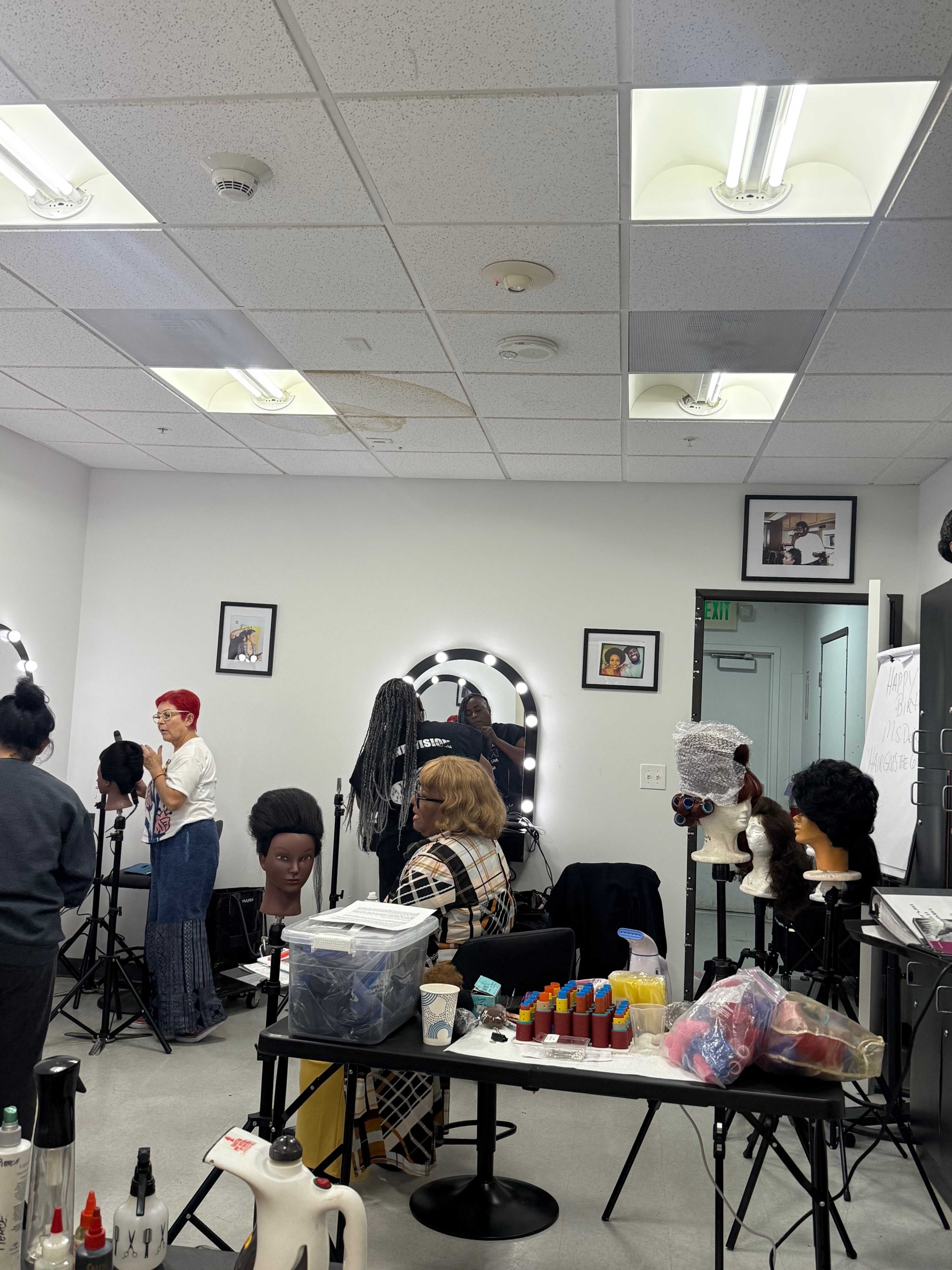 A cosmetology classroom features several students working on mannequins, with styling tools and various hair products displayed on a table.