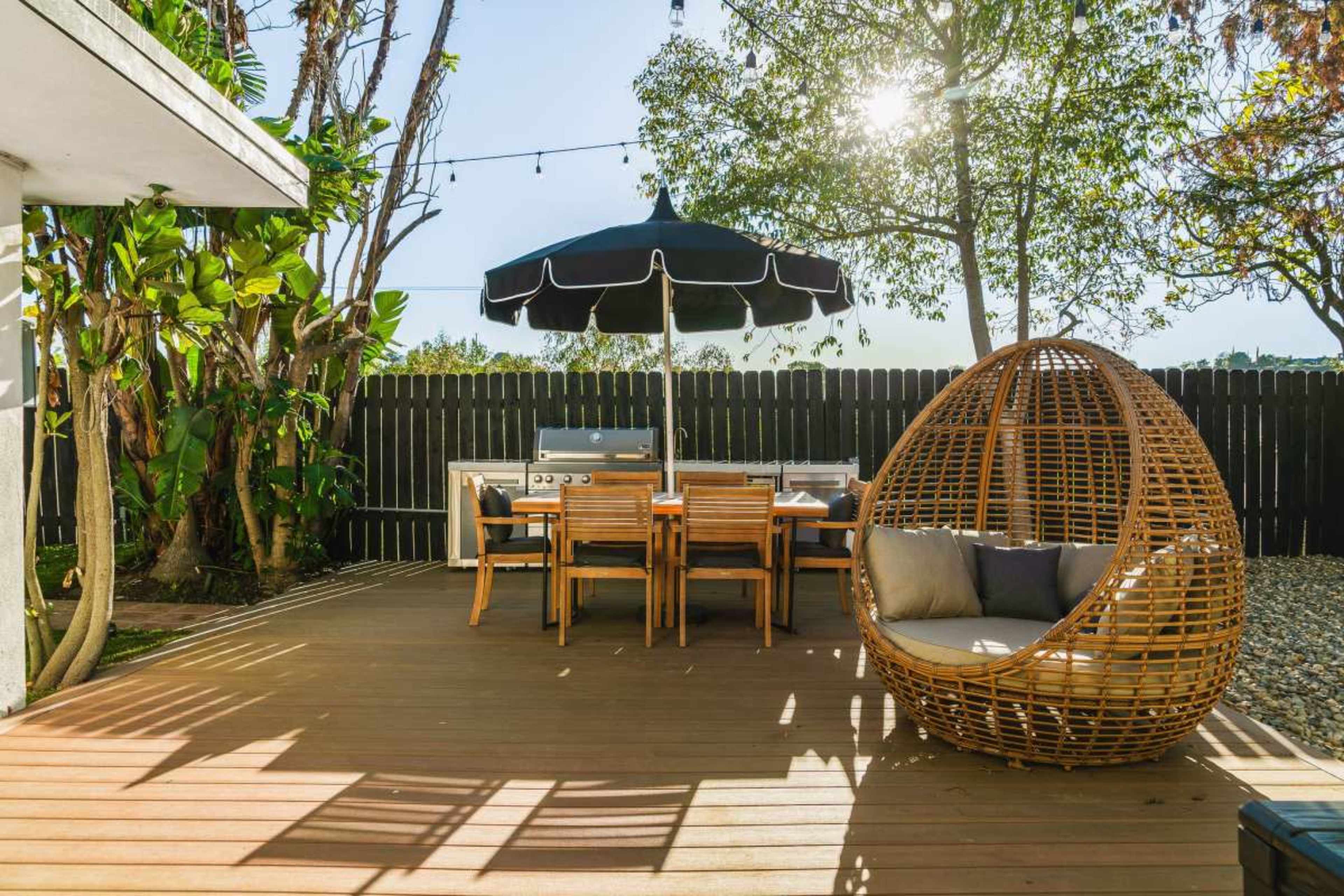 A wooden deck features a dining table with chairs, an umbrella, a grill, and a woven egg-shaped chair surrounded by greenery.