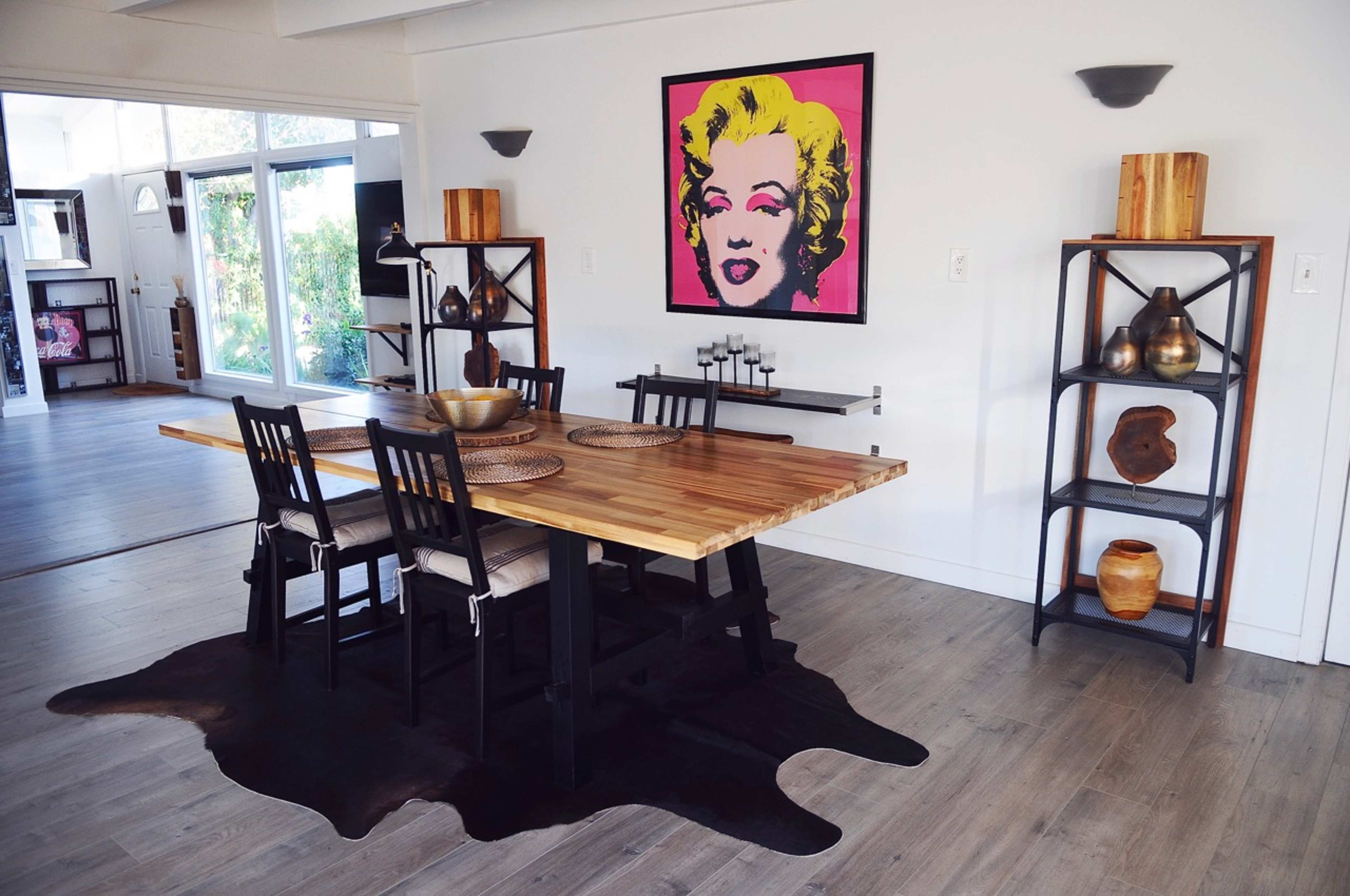 The image shows a dining area with a wooden table and black chairs on a dark rug, featuring an artwork of a woman on the wall and decorative shelving units.