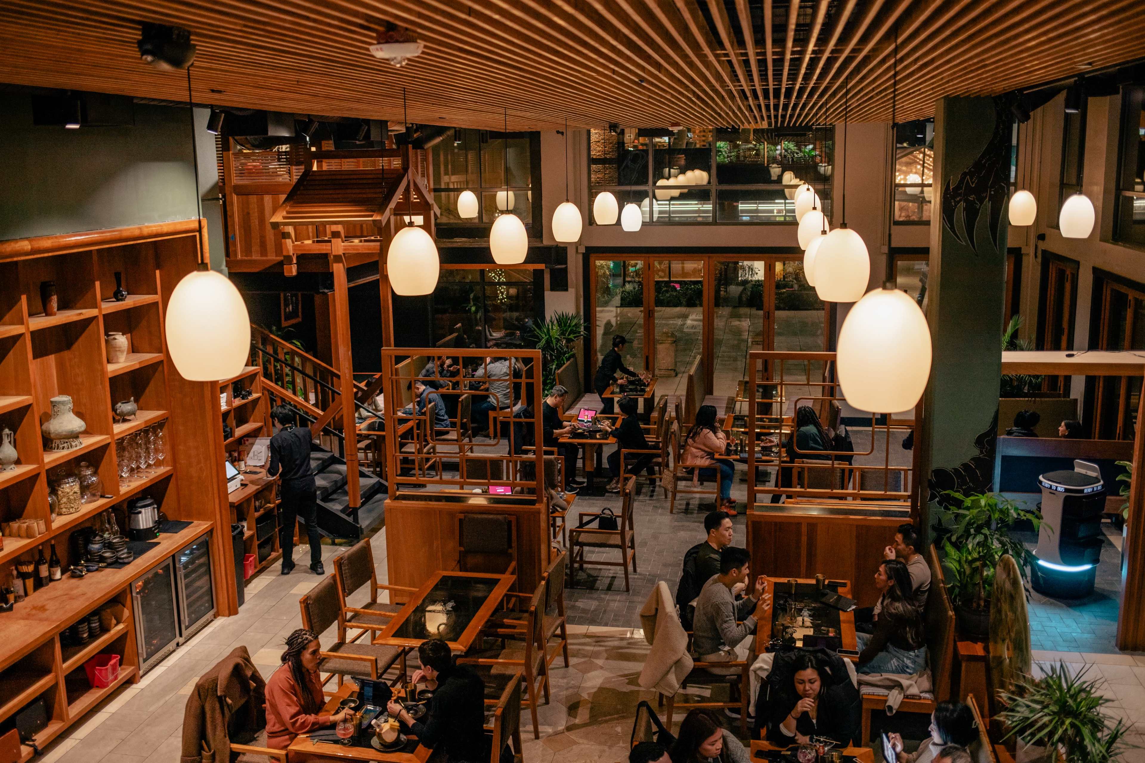 The image shows a spacious restaurant interior with wooden furnishings, softly lit pendant lamps, and patrons seated at tables engaged in conversation.