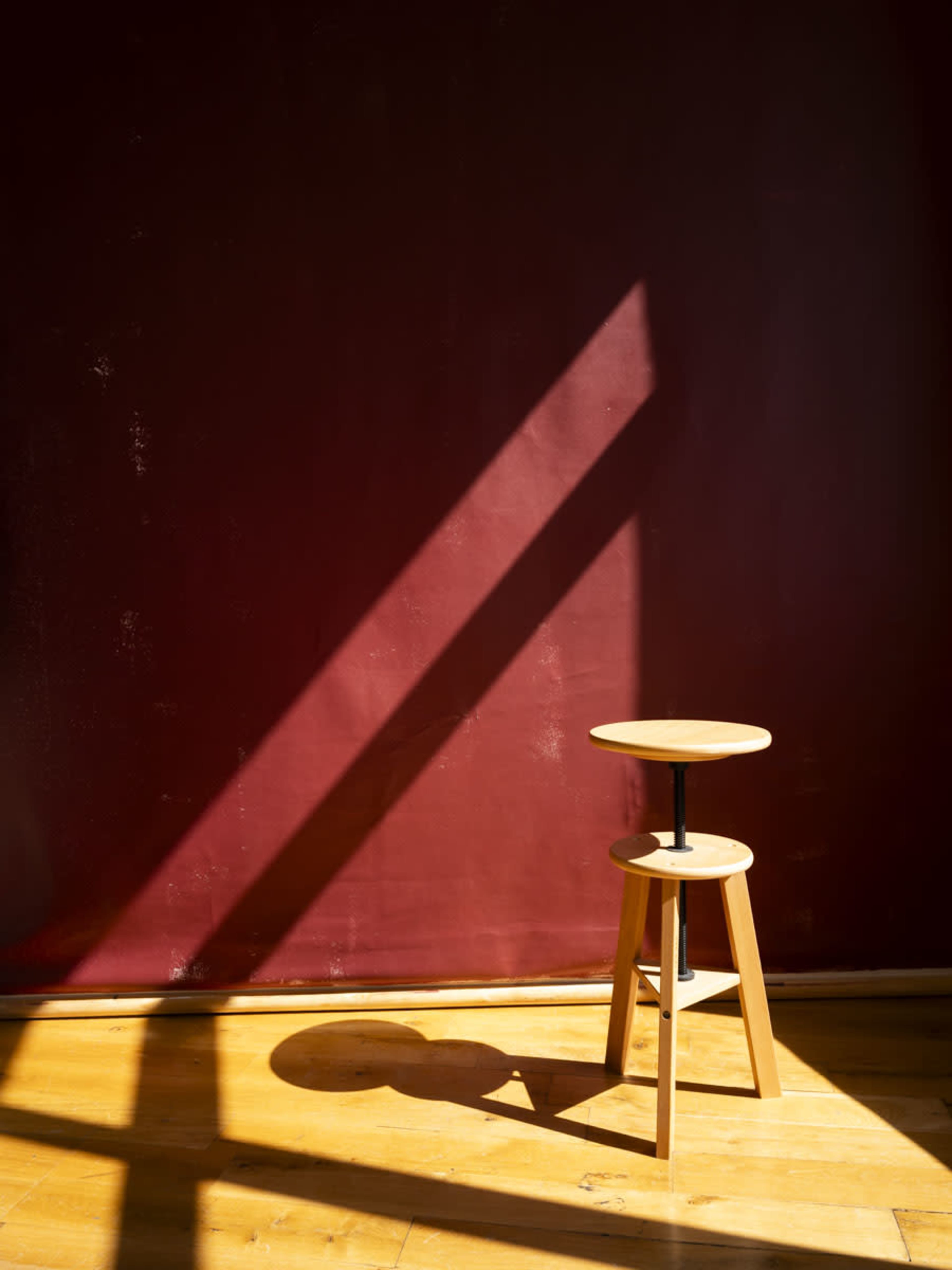 A wooden stool stands on a wooden floor, illuminated by sunlight casting a diagonal shadow against a dark red wall.