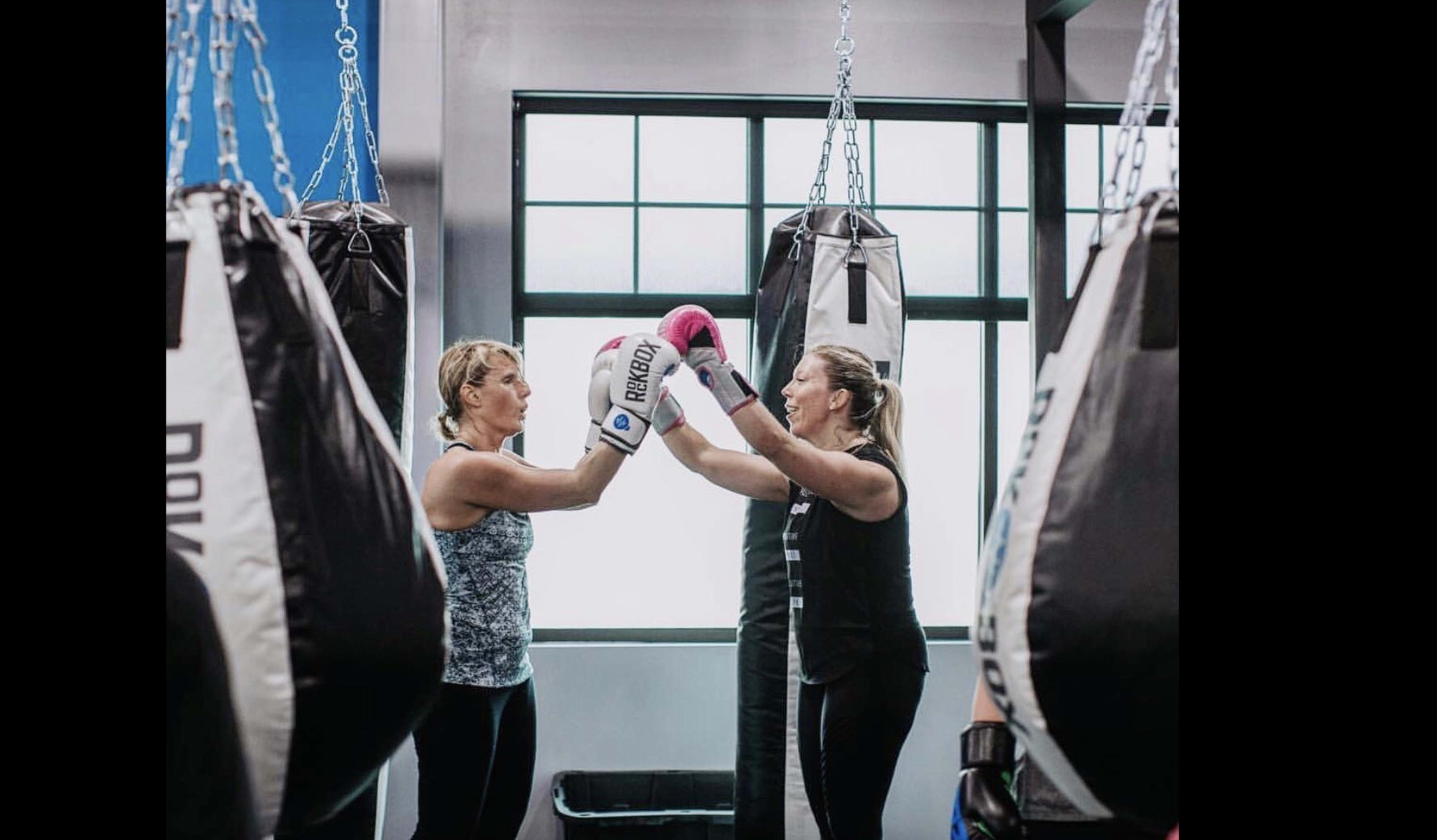 Two women are sparring in a boxing gym surrounded by hanging punching bags.