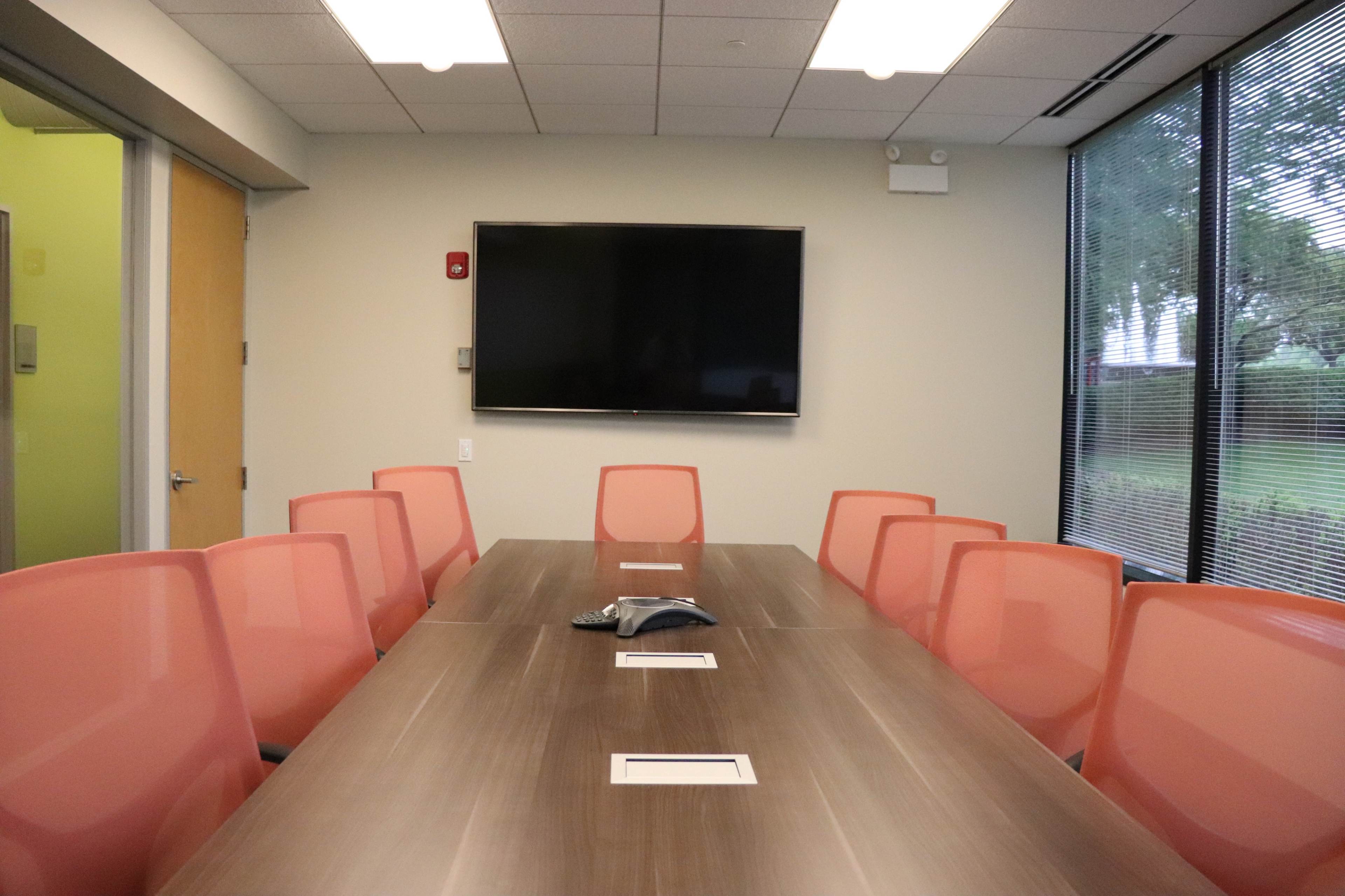 A conference room features a long wooden table surrounded by eight pink chairs, with a wall-mounted TV and large windows allowing natural light.