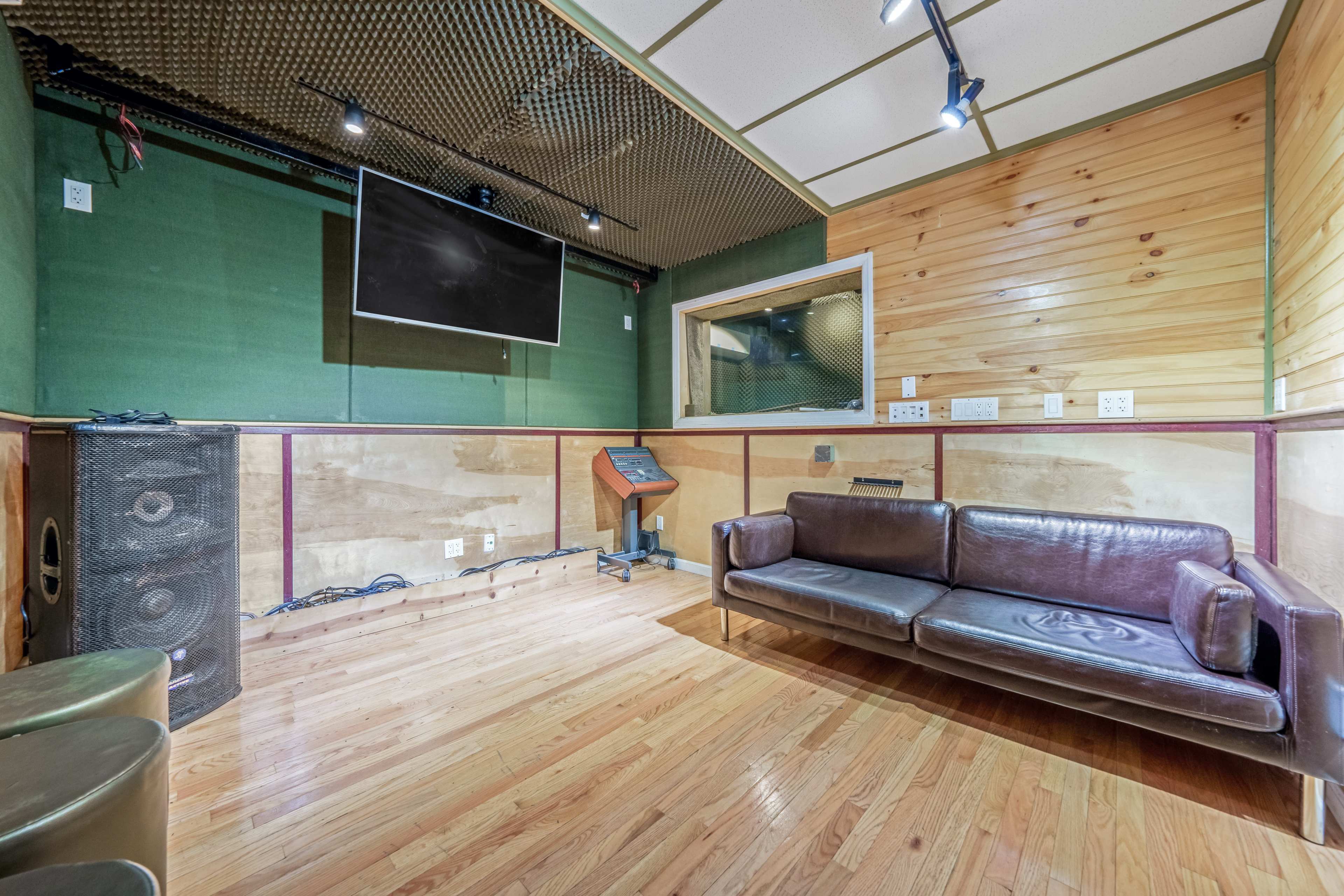 The image shows a sound recording studio with wooden walls, a television mounted on the wall, and a leather couch positioned on the floor.